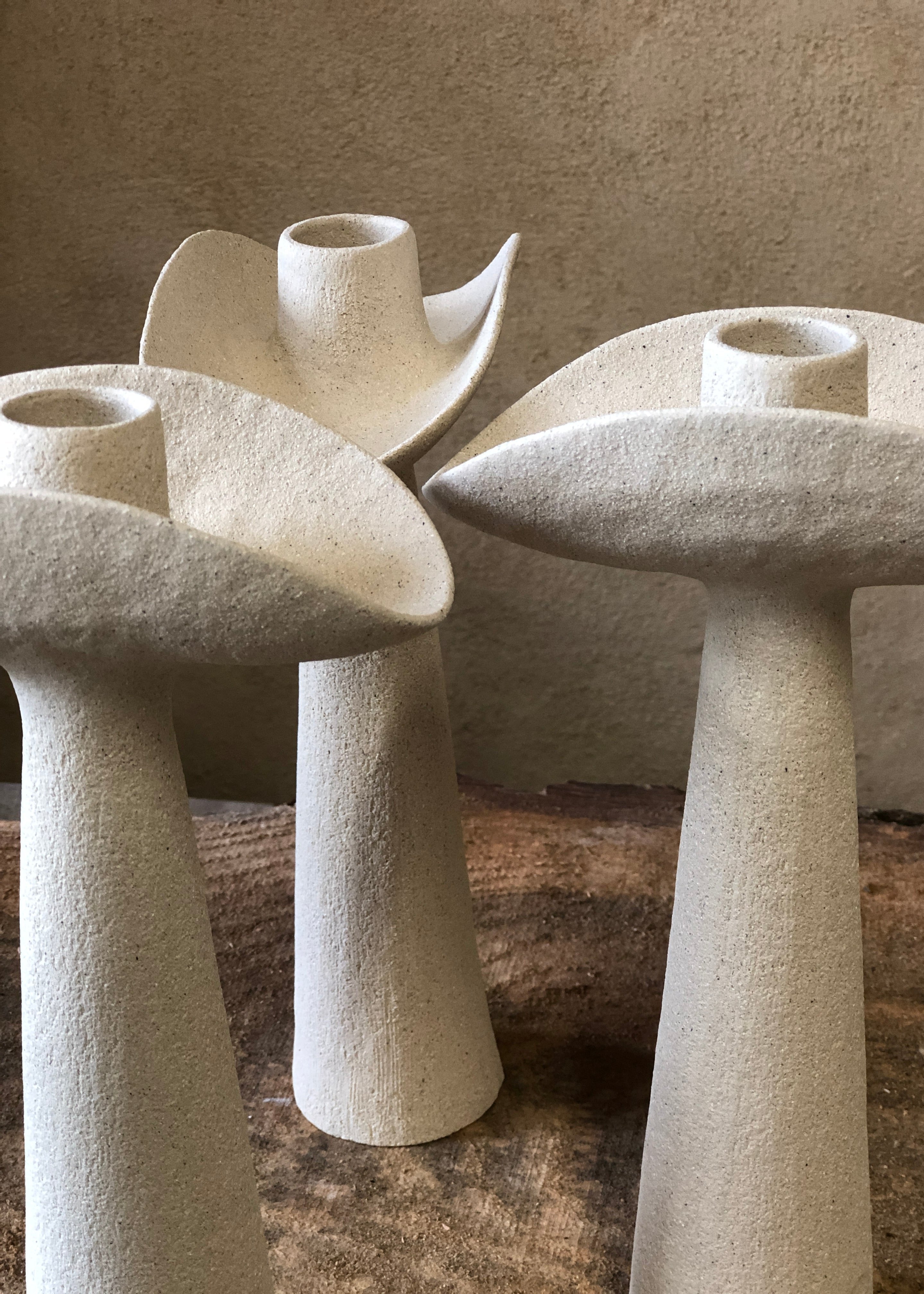 Bougeoirs en grès de design moderne, présentés en trio, avec des formes sculpturales. Les bougeoirs se distinguent par des bases coniques et des têtes évasées, créant un contraste entre leurs lignes douces et texturées. La surface présente une finition mate, mettant en valeur la couleur beige et la texture naturelle du grès, sur un fond neutre qui accentue leur élégance artisanale.