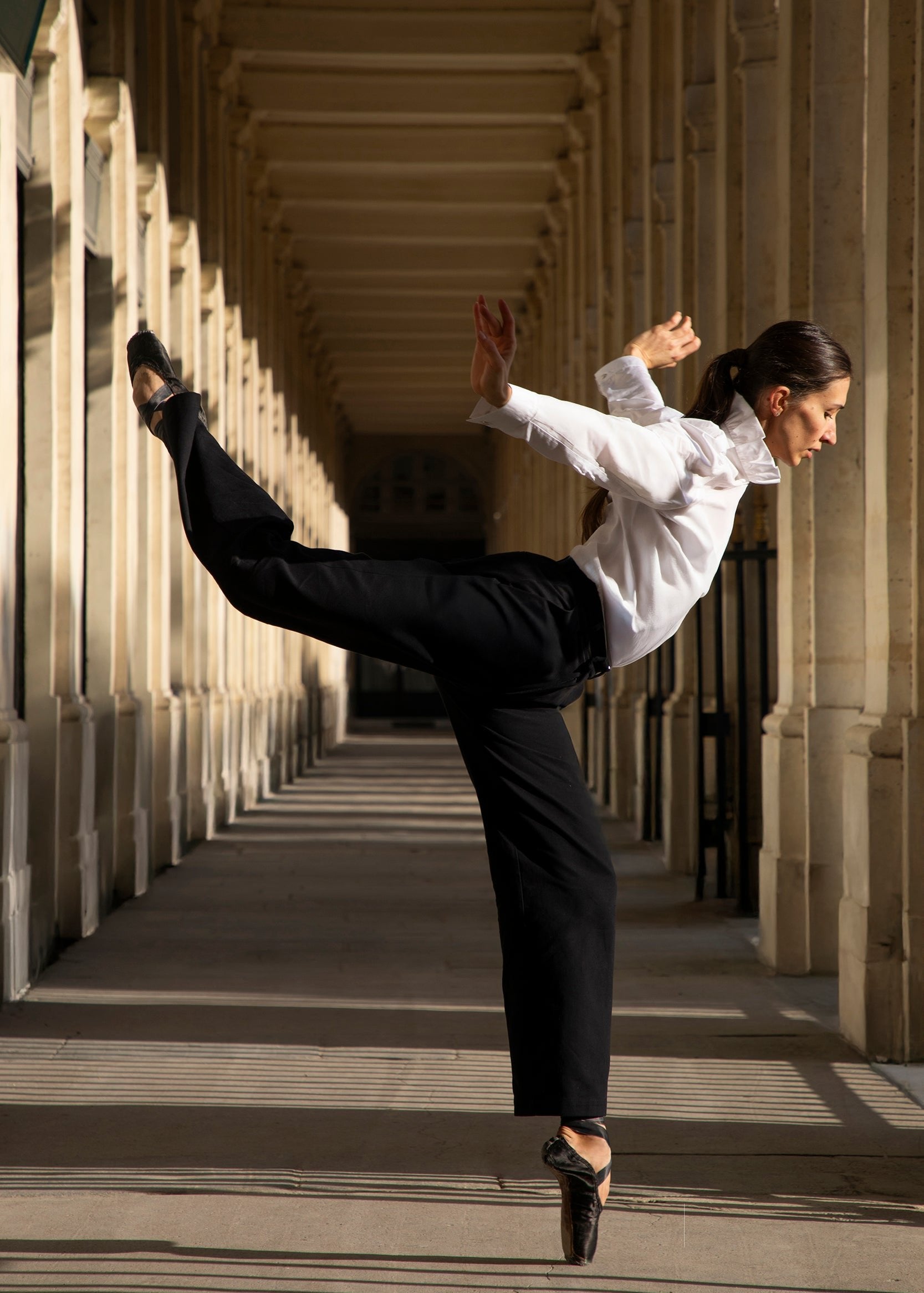 Une danseuse exécute avec élégance une gracieuse arabesque dans un couloir bordé de colonnes. Vêtue du Pantalon Raymonda de Sidonie Paris et d'un chemisier blanc, elle est en pointe, une jambe tendue derrière elle, projetant une ombre sur le sol. La scène est éclairée par la lumière naturelle.