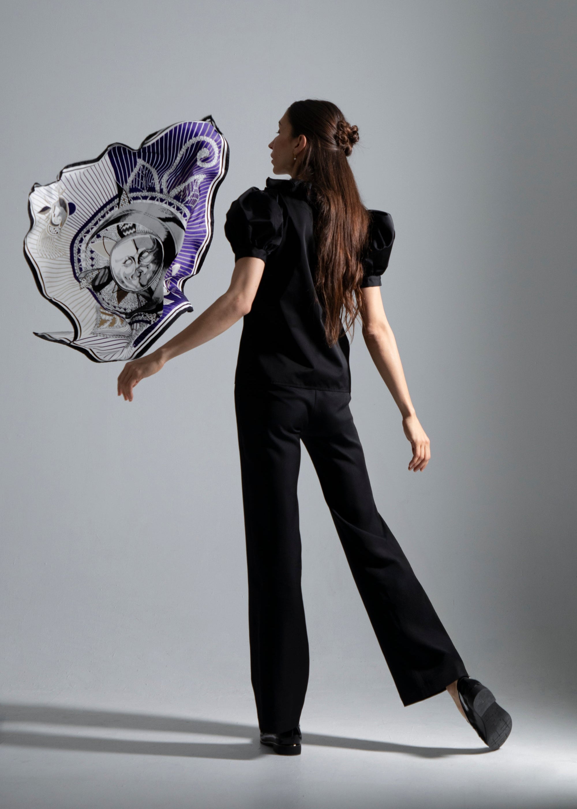 Foulard Ombre et Lumière flottant délicatement à côté d’une femme de dos, vêtue du top noir Vesper et du pantalon noir Salomé. Le foulard contraste un bleu indigo profond sur une moitié et un blanc lumineux sur l’autre, orné de rayons bleus et dorés. Au centre, une gravure noire et blanche représente le soleil et la lune, stylisés en forme de fleur évoquant un cadran brodé, avec deux motifs graphiques imaginaires le regardant.