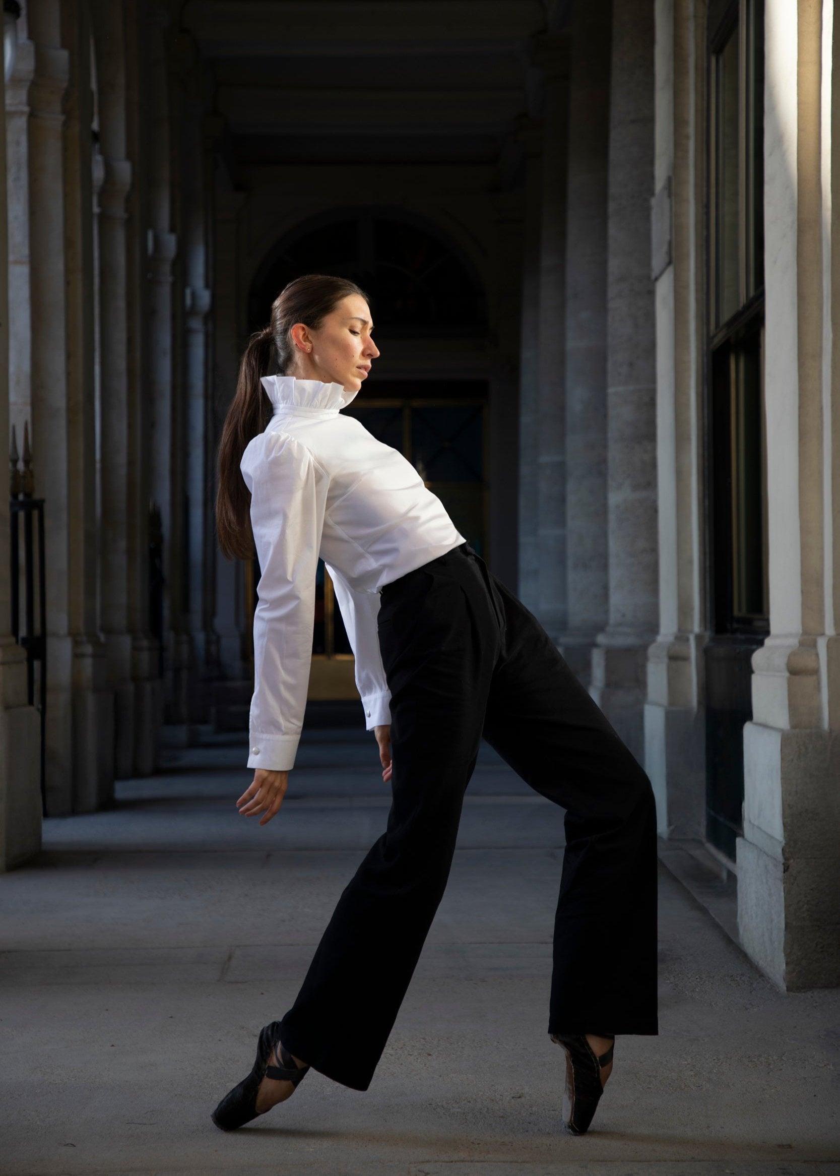 Dans un couloir aux hautes colonnes, une danseuse de ballet en blouse blanche et pantalon Raymonda prend une pose artistique, appuyée sur une pointe. La scène est éclairée de façon spectaculaire, soulignant l'élégance luxueuse de son mouvement gracieux.