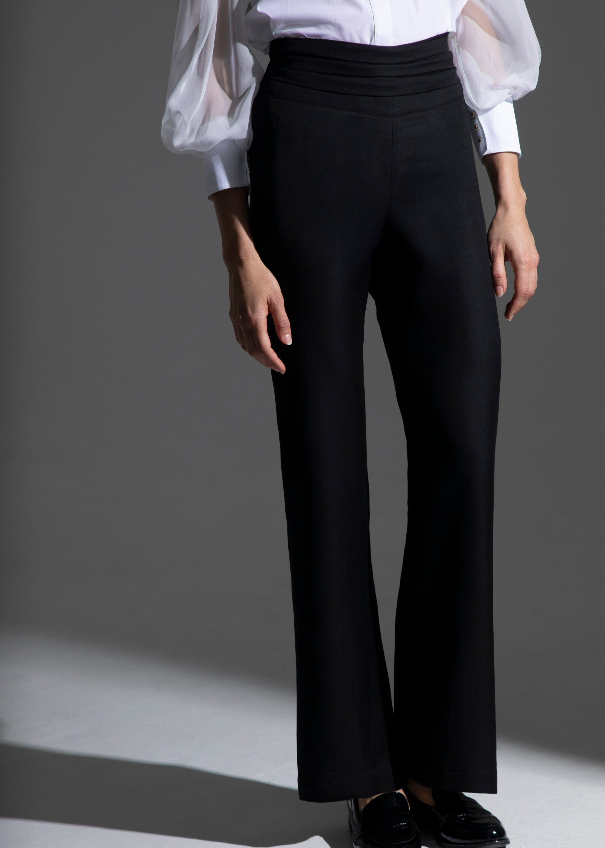 Femme portant le pantalon Salomé noir en laine et soie, souligné par une ceinture à plis horizontaux inspirée des pantalons de smoking masculins. Le jeu d’ombre et de lumière souligne la texture et la coupe élégante du vêtement.