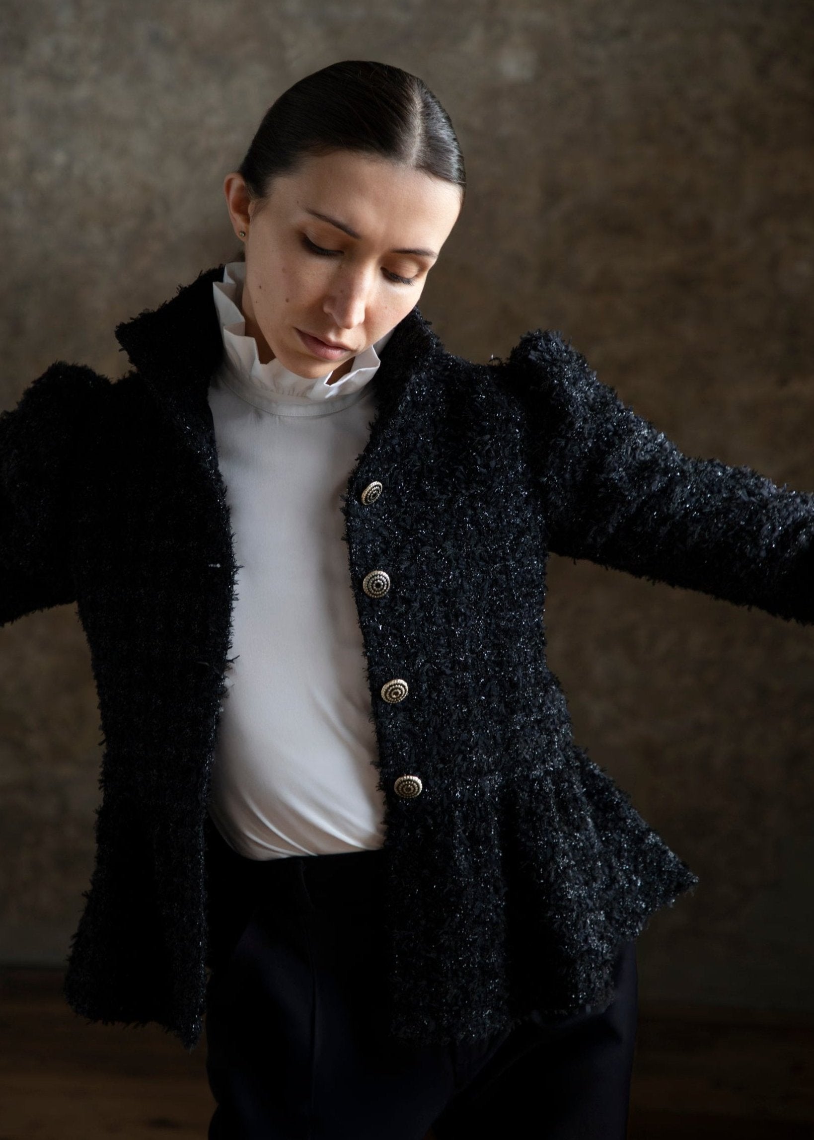Veste en tweed noir pailleté dotée de boutons en métal, portée par une femme qui bouge dans son vêtement. La veste présente des manches bouffantes et un col montant, tandis qu'elle porte un chemisier blanc en dessous. L'arrière-plan est flou, mettant en valeur le style sophistiqué et élégant de la tenue.