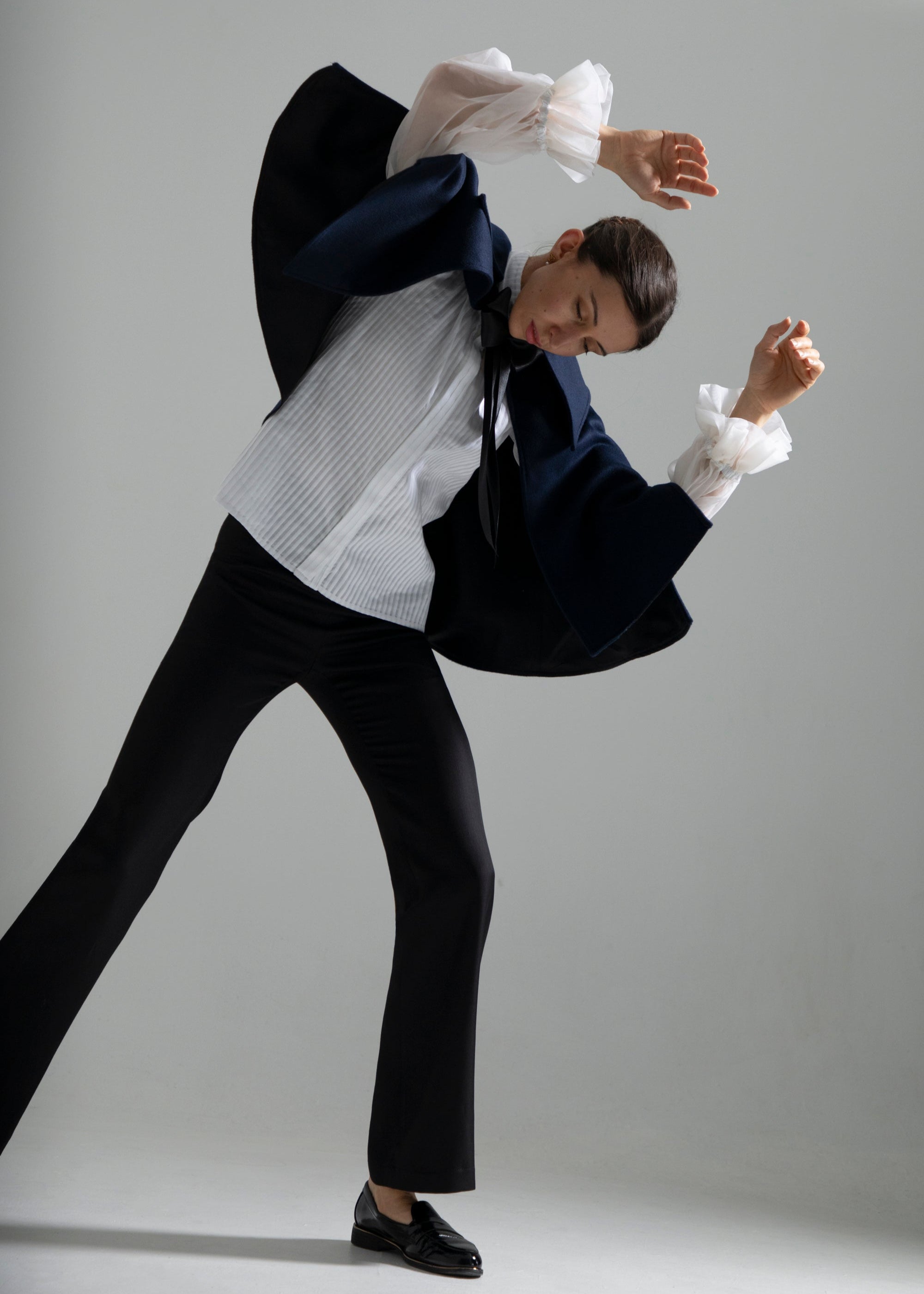 Femme vue de face, se balançant avec grâce, les deux bras levés en l’air et le regard dirigé vers le sol. Elle porte la cape Ondine bleu nuit, en laine et soie, courte à la taille, avec des fentes avant laissant passer les bras librement. La doublure noire de la cape est partiellement visible. Sous la cape, le chemisier Isolde blanc et le pantalon Salomé noir complètent la tenue.