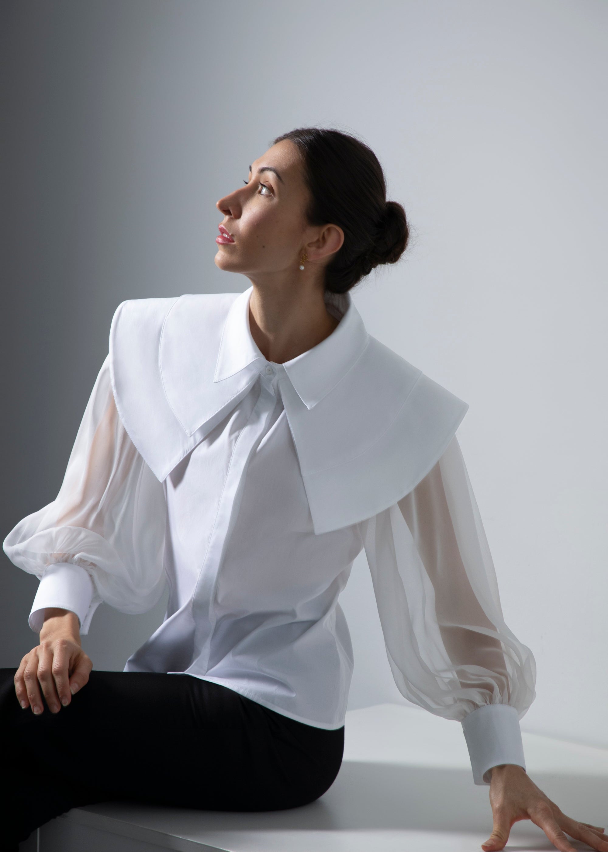 Femme assise, le regard tourné vers la lumière, portant le chemisier Albane à triple col, avec manches en organza de soie et poignets fermés par trois boutons dorés en forme de boule.
