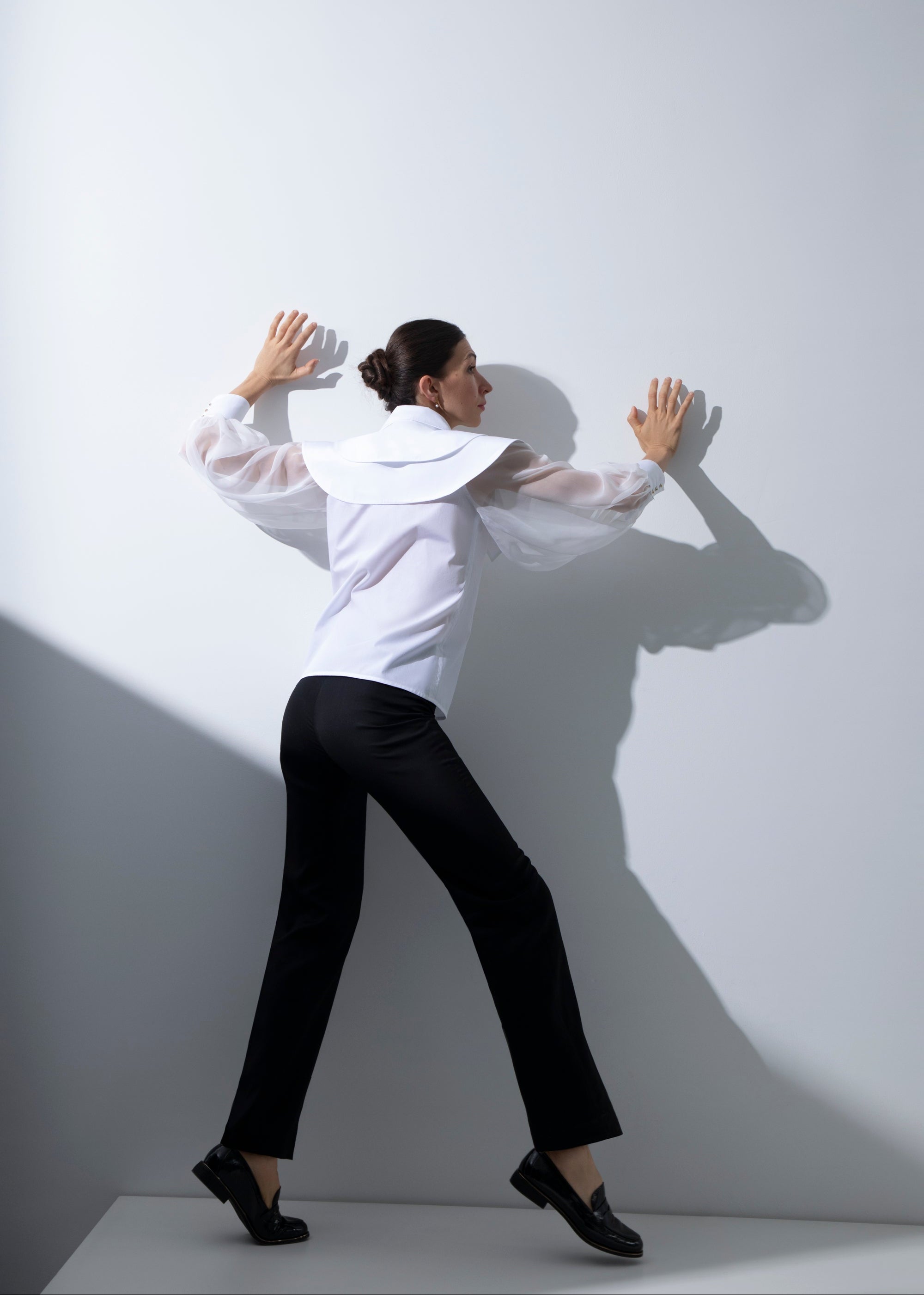 Femme de dos, face au mur, les mains posées à plat contre celui-ci. Elle porte le chemisier Albane, laissant apparaître la transparence de ses manches en organza de soie, assorti au pantalon noir Salomé.