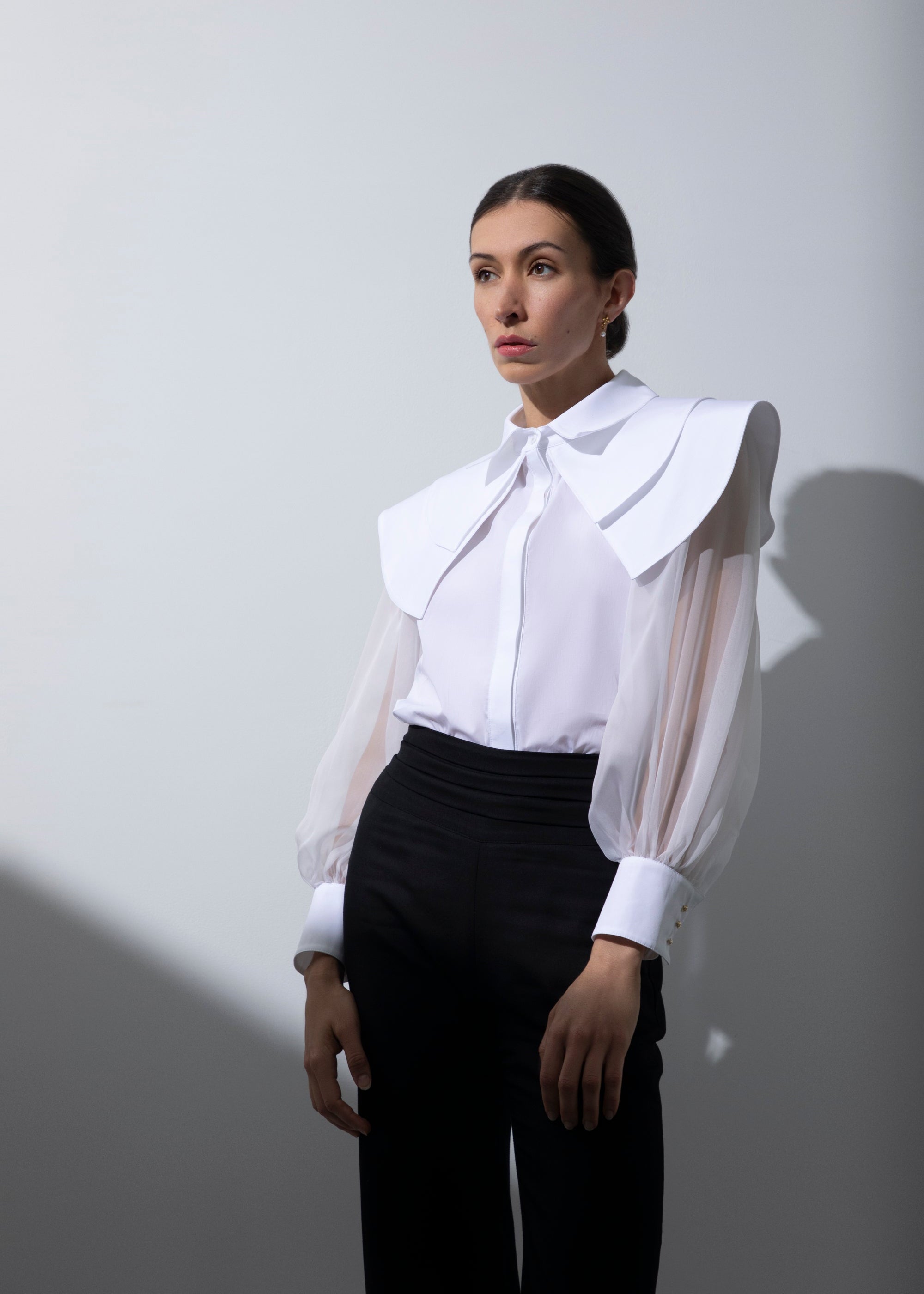 Femme debout, de face, se tenant droite, vêtue du chemisier Albane à triple col et du pantalon noir Salomé. La transparence des manches en organza de soie contraste avec la structure épurée de la silhouette.