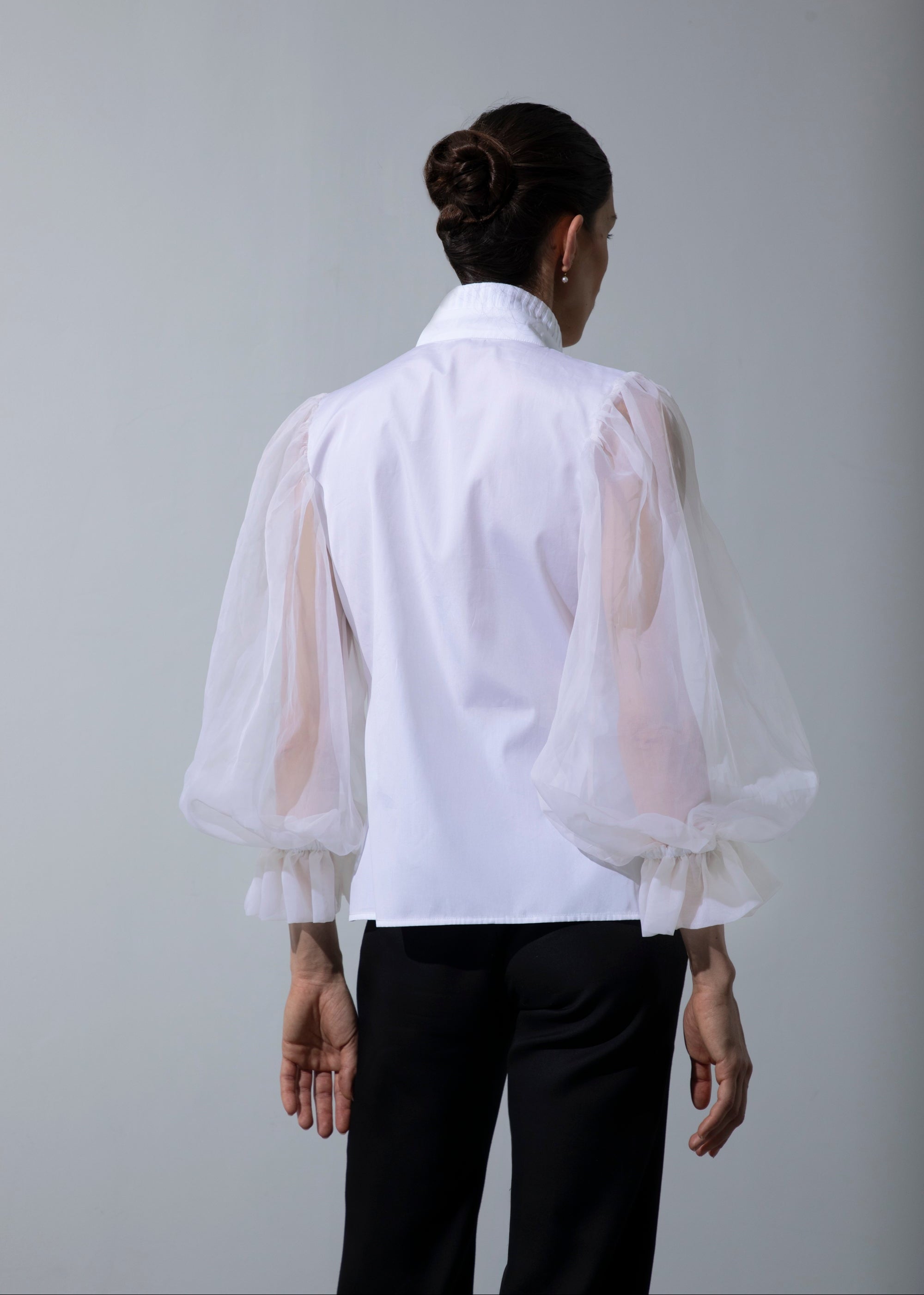 Une femme se tient debout, de dos, et porte le chemisier Isolde, tout en contraste, avec ses manches transparentes et son dos opaque blancs, assorti au pantalon noir Salomé.