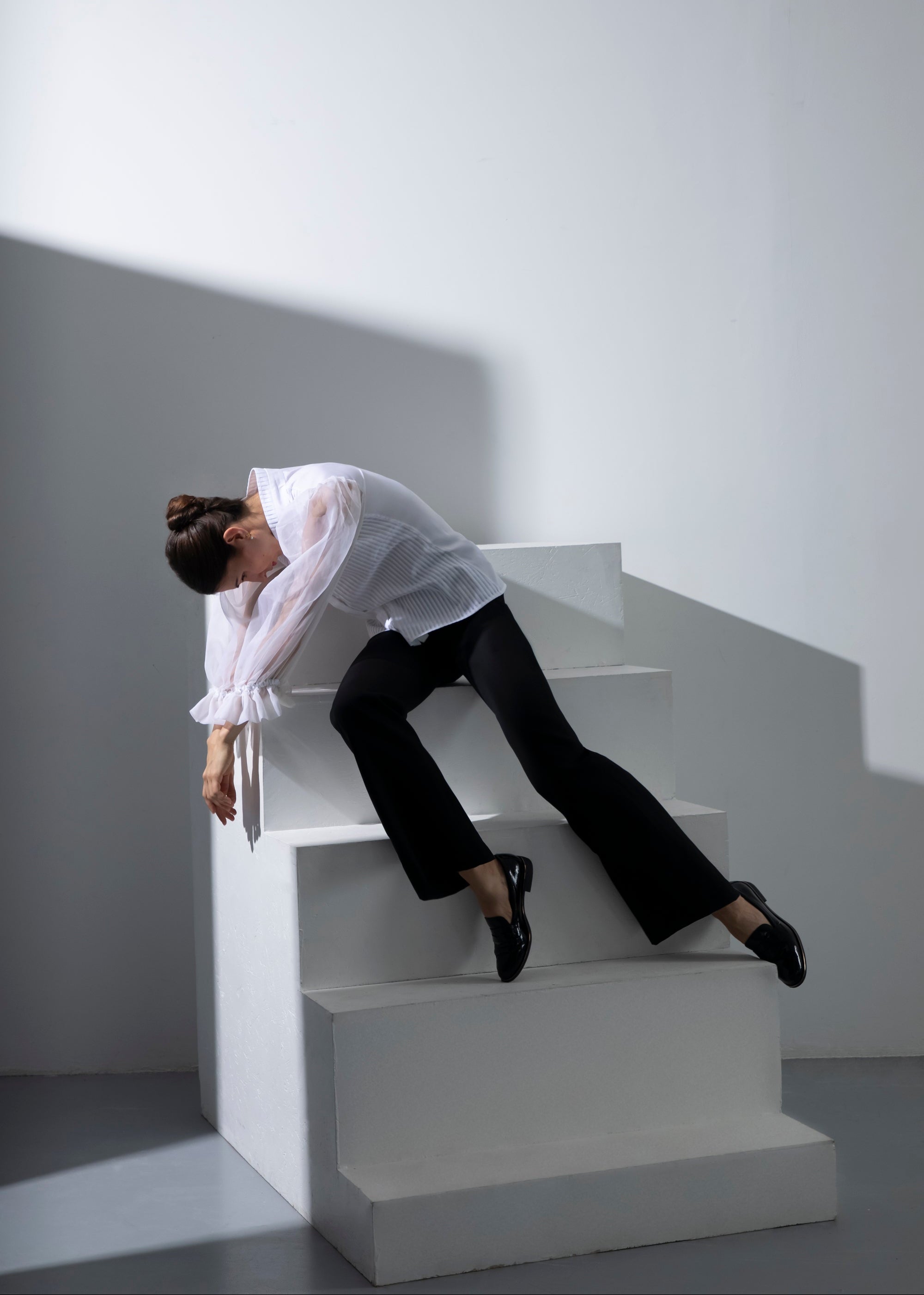 Une femme assise se penche sur un escalier blanc, vêtue d'un chemisier Isolde blanc et d'un pantalon Salomé noir, jouant avec ombre et lumière.