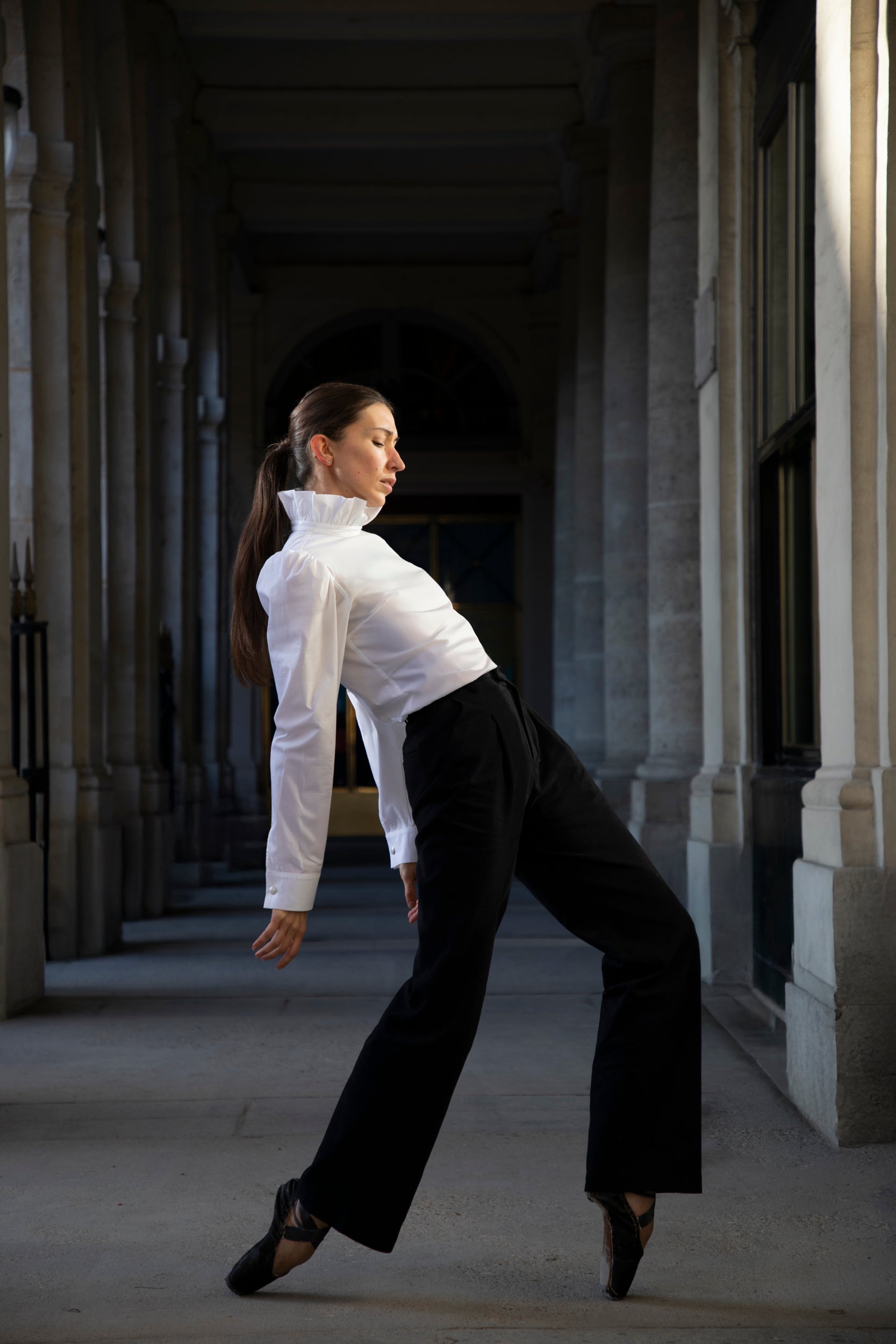 Une danseuse vêtue du Pantalon Raymonda de Sidonie Paris se penche gracieusement en arrière dans un couloir bordé de colonnes de pierre. L'éclairage met élégamment en valeur sa pose et les détails architecturaux environnants, créant une aura de luxe.