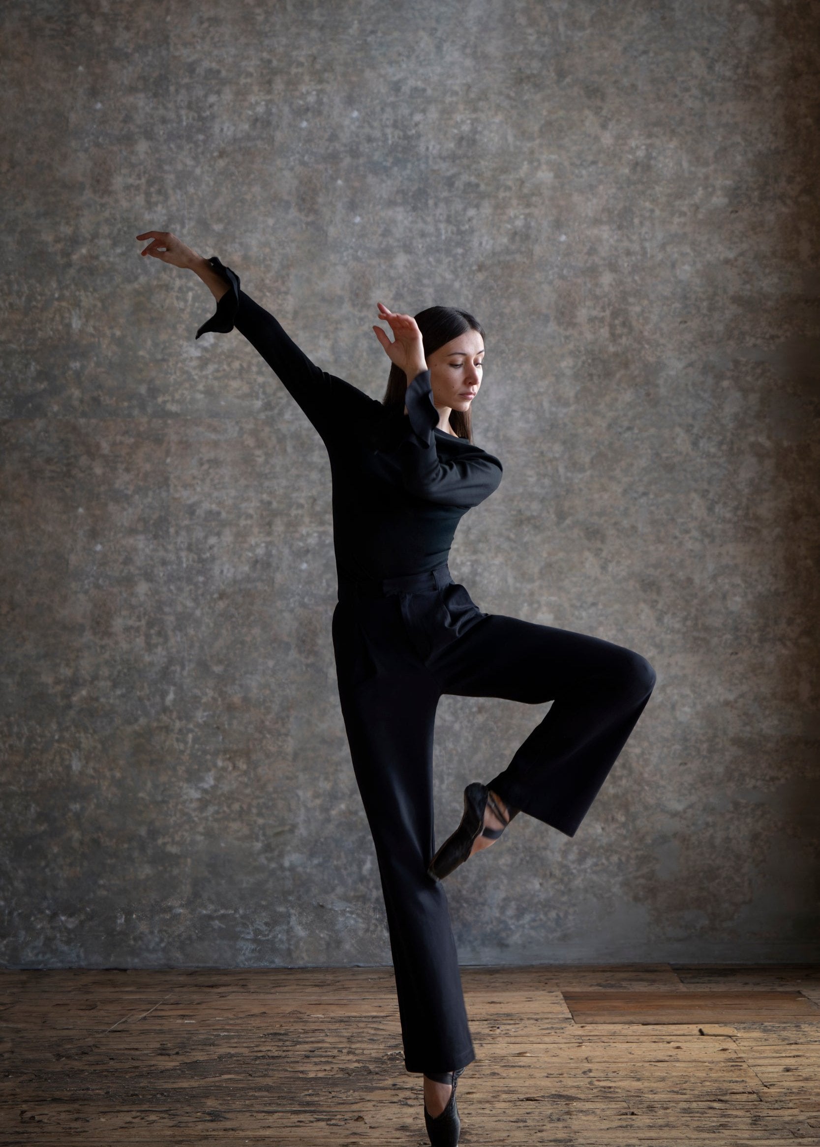 Une danseuse de ballet exécute une pose artistique élégante, vêtue d'un t-shirt à volants et pantalon noir, sur un fond de mur texturé gris. Son bras est étendu gracieusement, tandis que sa jambe est levée dans une position dynamique, illustrant la beauté et la précision du ballet. L'image capture l'essence de l'art du mouvement, mettant en valeur le corps de la danseuse comme une véritable œuvre d'art en action.