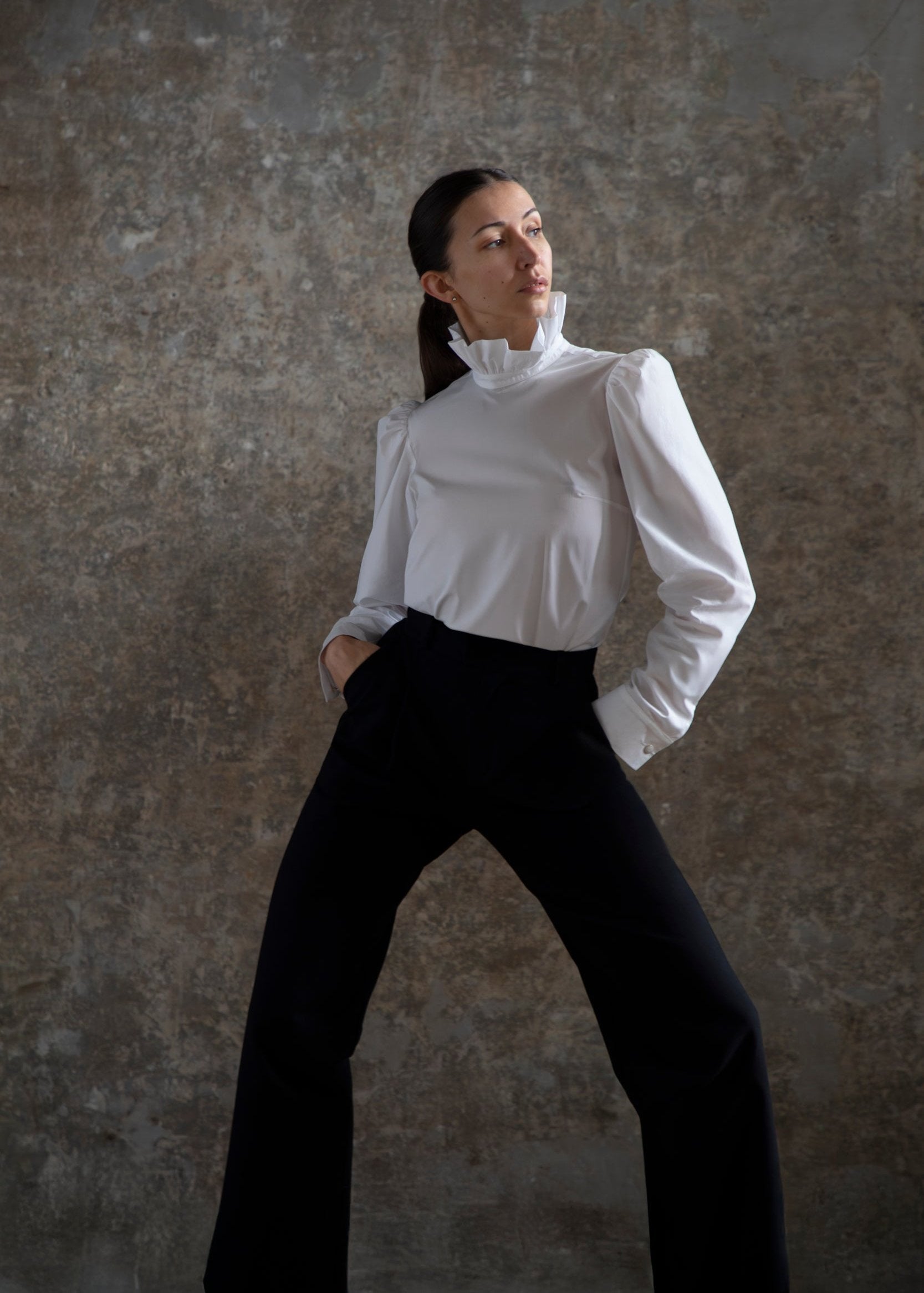 Sur un fond gris texturé, une femme respire l'élégance en portant le chemisier Sidonie. Une chemise blanche à col montant, associée à un pantalon noir. Elle pose avec assurance, les mains dans les poches, les jambes écartées et le regard légèrement vers le haut, incarnant parfaitement l'essence de l'art moderne.