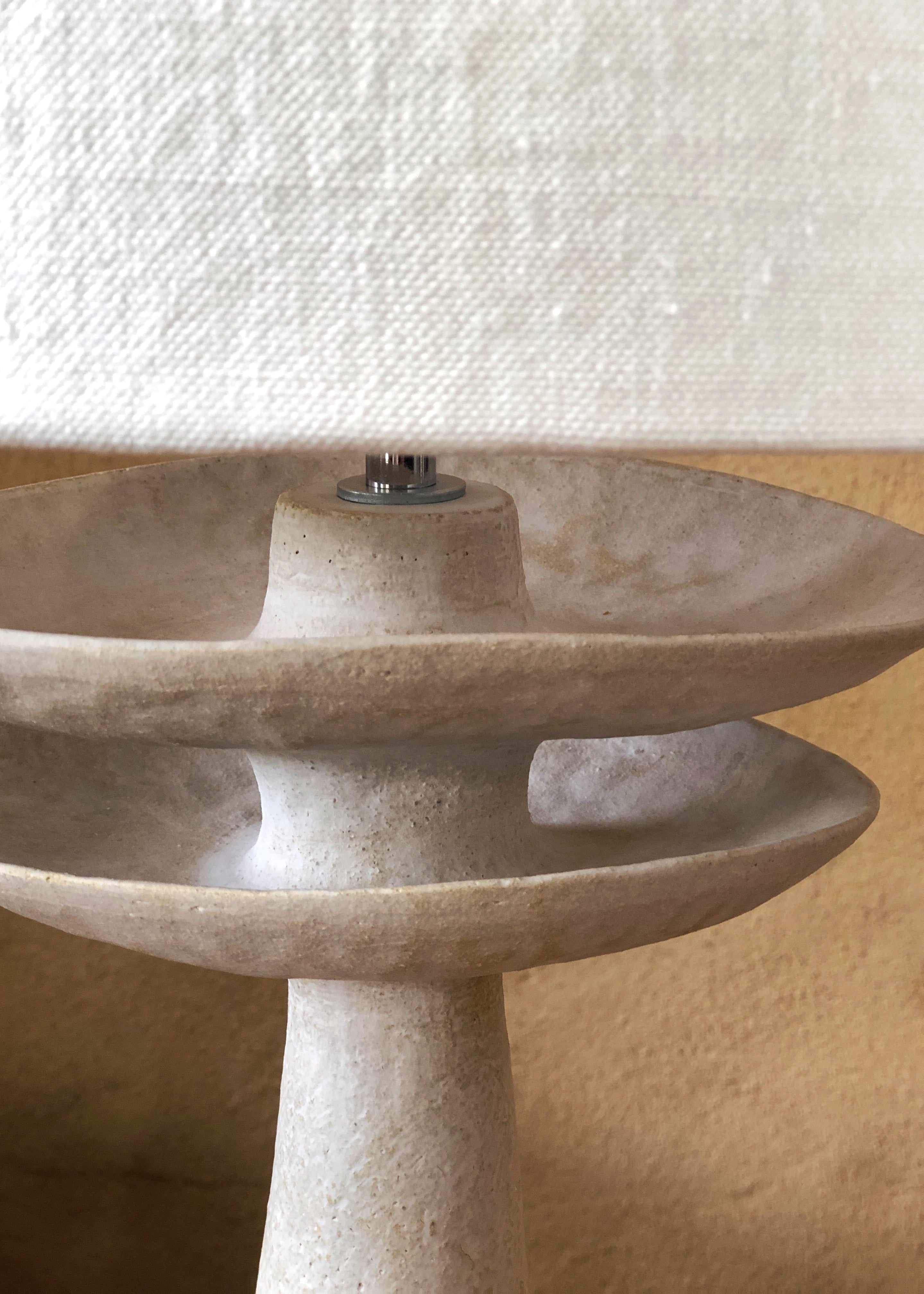 Lampe en grès beige avec un design élégant, présentant une base en forme de colonne et deux niveaux de platines. La partie supérieure est recouverte d'un abat-jour en lin naturel, créant une atmosphère douce. La texture de la céramique est visible, ajoutant un élément artisanal à l'objet. Le tout repose sur un fond neutre, accentuant la couleur beige et le style minimaliste de cette lampe décorative.