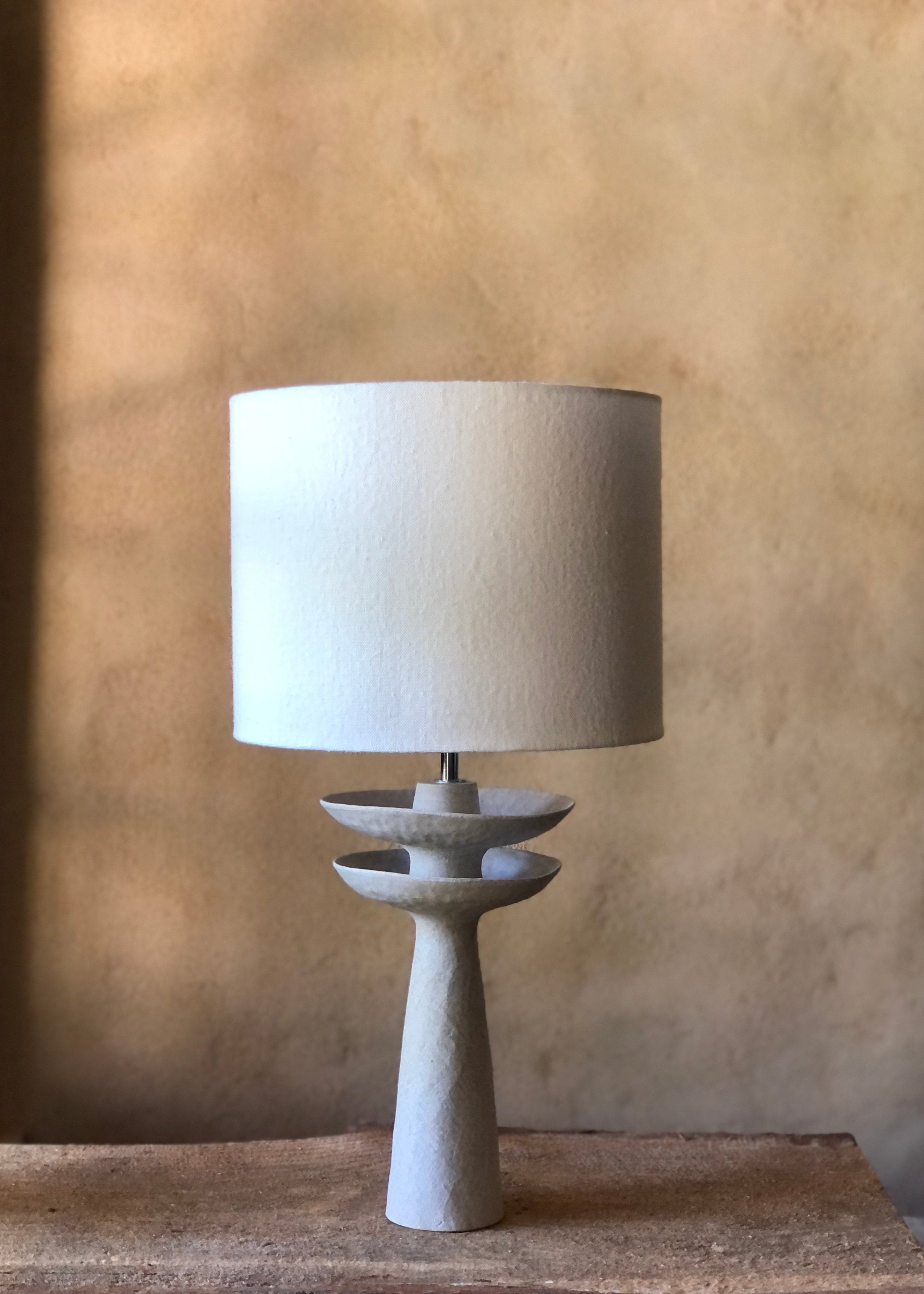 Lampe moderne en grès beige avec un abat-jour cylindrique en lin naturel, reposant sur une base sculpturale à deux étages circulaires. Cette lampe, objet d'art, présente des couleurs neutres et des formes épurées, créant une ambiance élégante et contemporaine. Elle est placée sur une surface en bois texturé, avec un fond neutre qui met en valeur son design minimaliste.