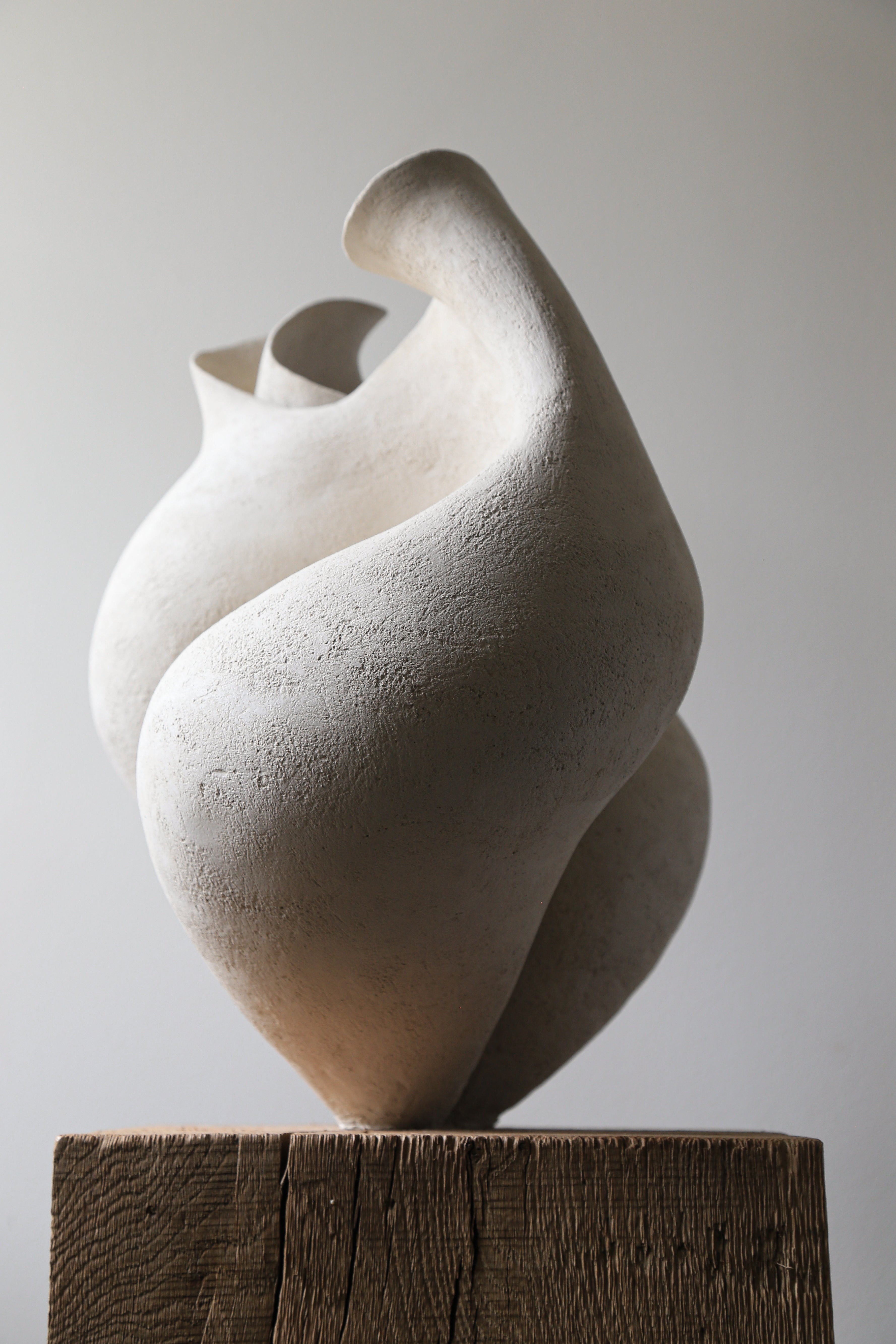 Sculpture moderne en grès écru, présentant des formes organiques et fluides. La pièce se dresse sur un socle en bois brut, mettant en valeur sa surface texturée et ses courbes élégantes. Les nuances de couleur et la lumière jouent sur sa surface, créant un effet visuel captivant qui attire l'œil. L'arrière-plan neutre accentue la beauté minimaliste de l'œuvre.