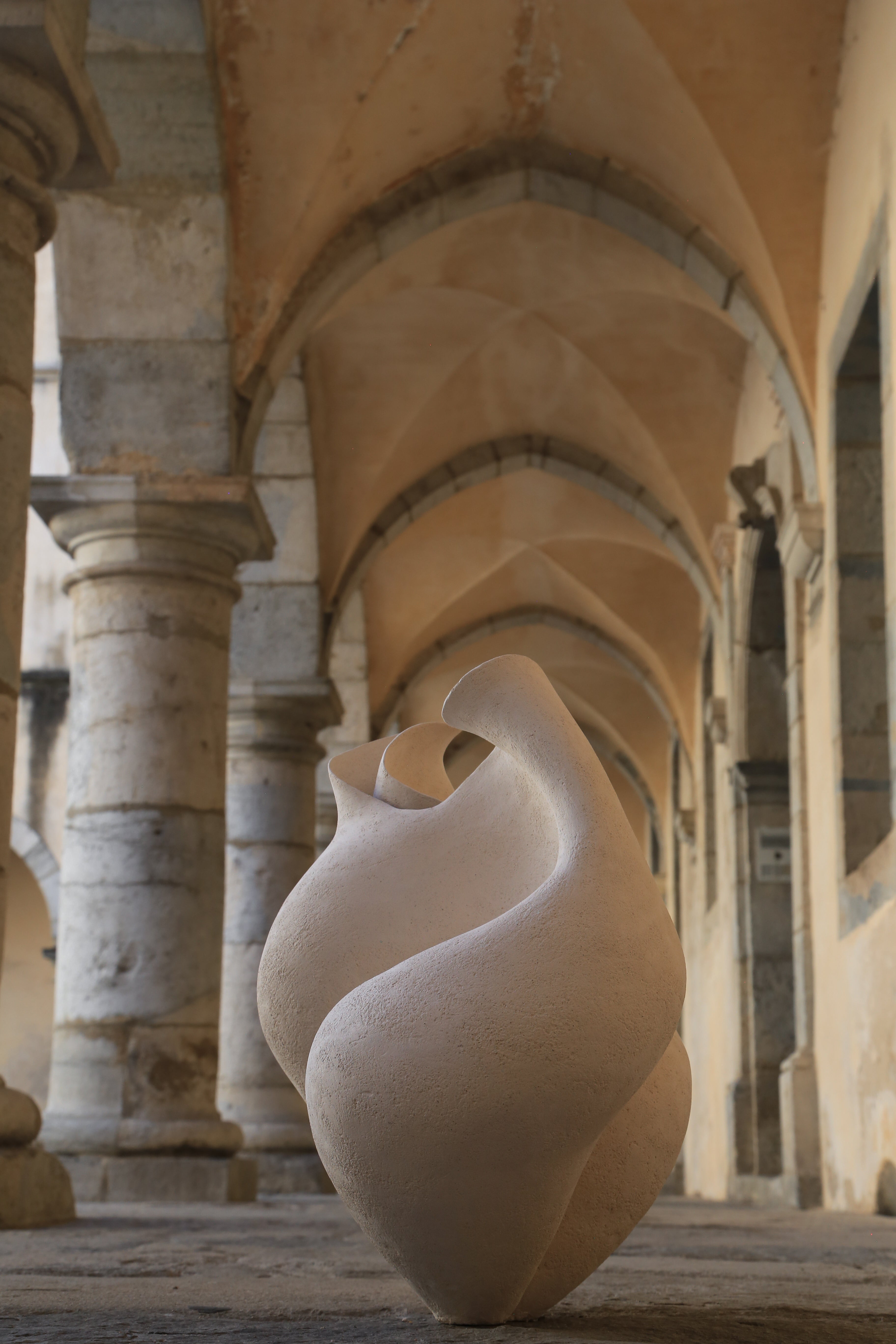 Sculpture en grès écru, au design fluide et organique, placée au centre d'un couloir voûté avec des arches en pierre. L'arrière-plan montre des murs aux teintes beige et des colonnes en pierre, créant une ambiance artistique et élégante. Cette œuvre d'art moderne évoque la créativité et l'harmonie, soulignant le contraste entre la douceur de la sculpture et la robustesse des éléments architecturaux environnants.