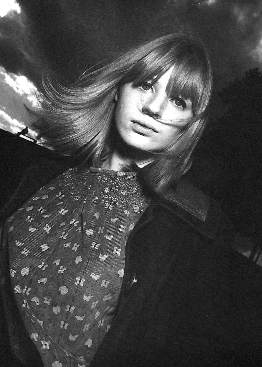 Page ouverte du livre Appearances de Martin Harrison montrant un portrait en noir et blanc,en pleine page, de Marianne Faithfull par David Bailey. Elle a des cheveux clairs au carré et avec une frange qui bouge avec le vent, elle se tient debout à l'extérieur avec derrière elle des arbres et des nuages gris. Elle porte une blouse et un manteau noir ouvert.