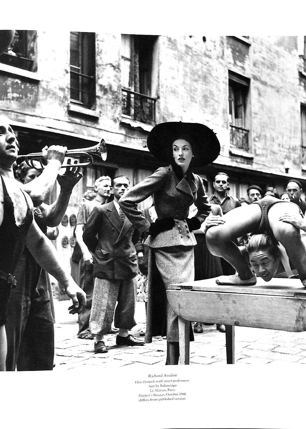 Page ouverte du livre Appearances de de Martin Harrison montrant une photo en noir et blanc d'une femme très élégante portant un tailleur Balenciaga, dans la rue au milieu d'une troupe d'artistes de rue et de spectateurs.