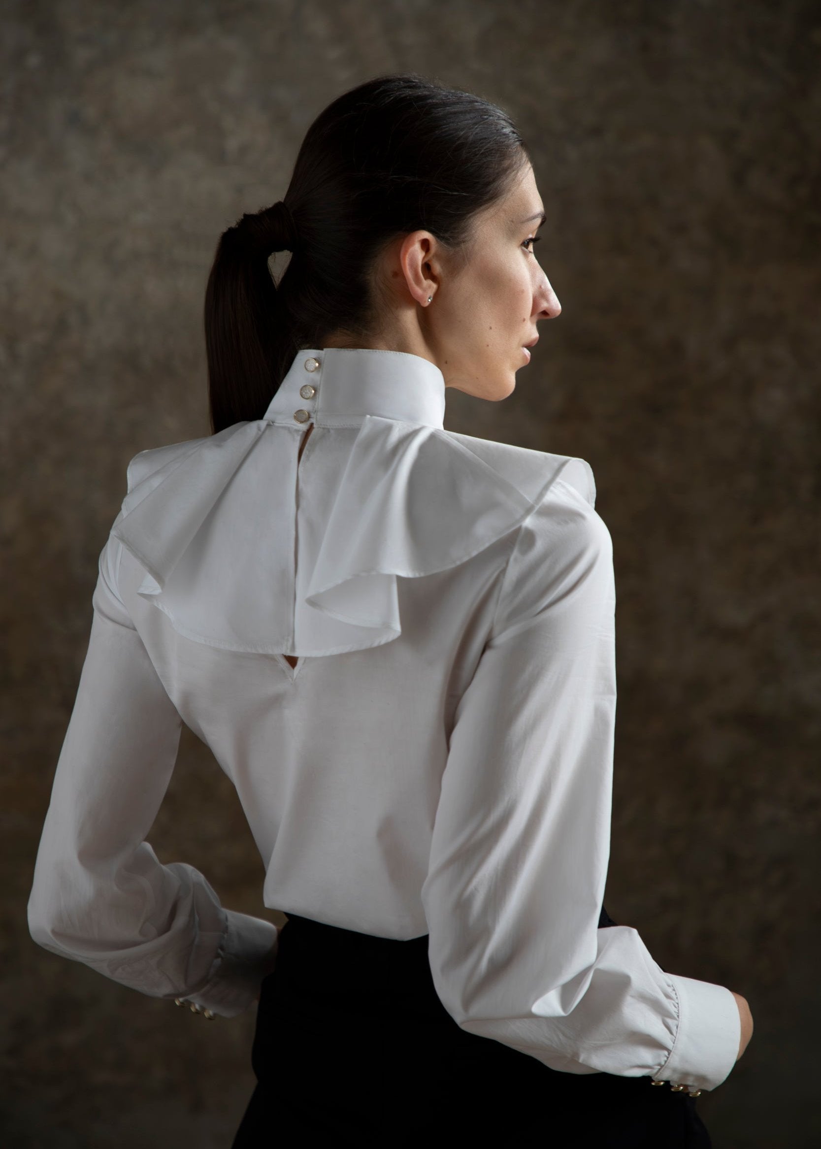 Une femme modèle porte une blouse blanche élégante Giselle, mettant en valeur un design sophistiqué avec des volants au dos. Les boutons dorés et nacrés se distinguent sur le col et les manches, ajoutant une touche de luxe à l'ensemble. La silhouette évoque le mouvement et la grâce du ballet, soulignant l'harmonie entre mode et art. Le fond flou met en valeur la beauté et l'élégance de la pièce.