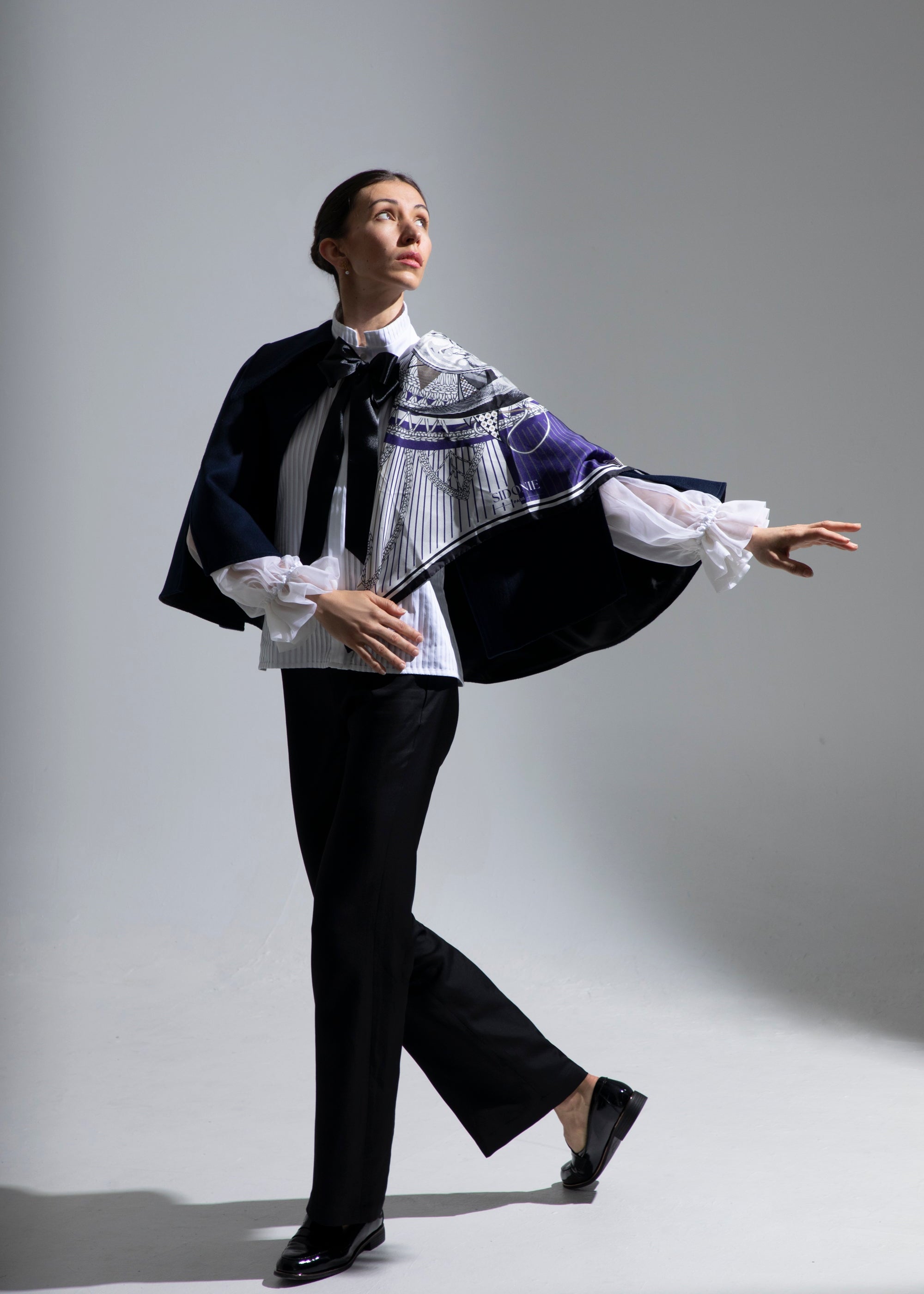 Femme marchant, vêtue de la cape Ondine, du chemisier Isolde et du pantalon Salomé, portant élégamment le foulard en soie Ombre et Lumière drapé sur le côté gauche de son haut du corps. Elle tend le bras gauche vers l’extérieur avec grâce, le regard tourné vers la lumière, capturant un moment de mouvement et de douceur.