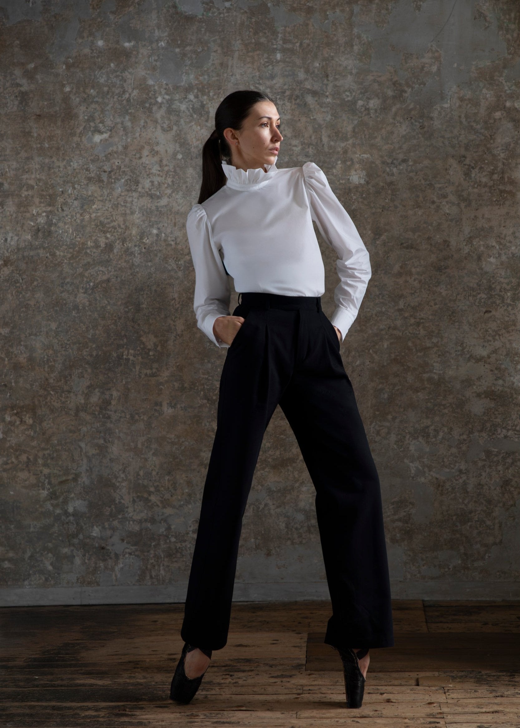 Une femme respire l'élégance, debout avec assurance sur des pointes de ballet noires, les mains dans les poches, vêtue d'un chemisier blanc à col montant associé au pantalon Raymonda, caractérisé par sa coupe large noire sophistiquée. Ses cheveux sont attachés en arrière sur un fond texturé aux tons neutres, créant une scène rappelant une œuvre d'art.