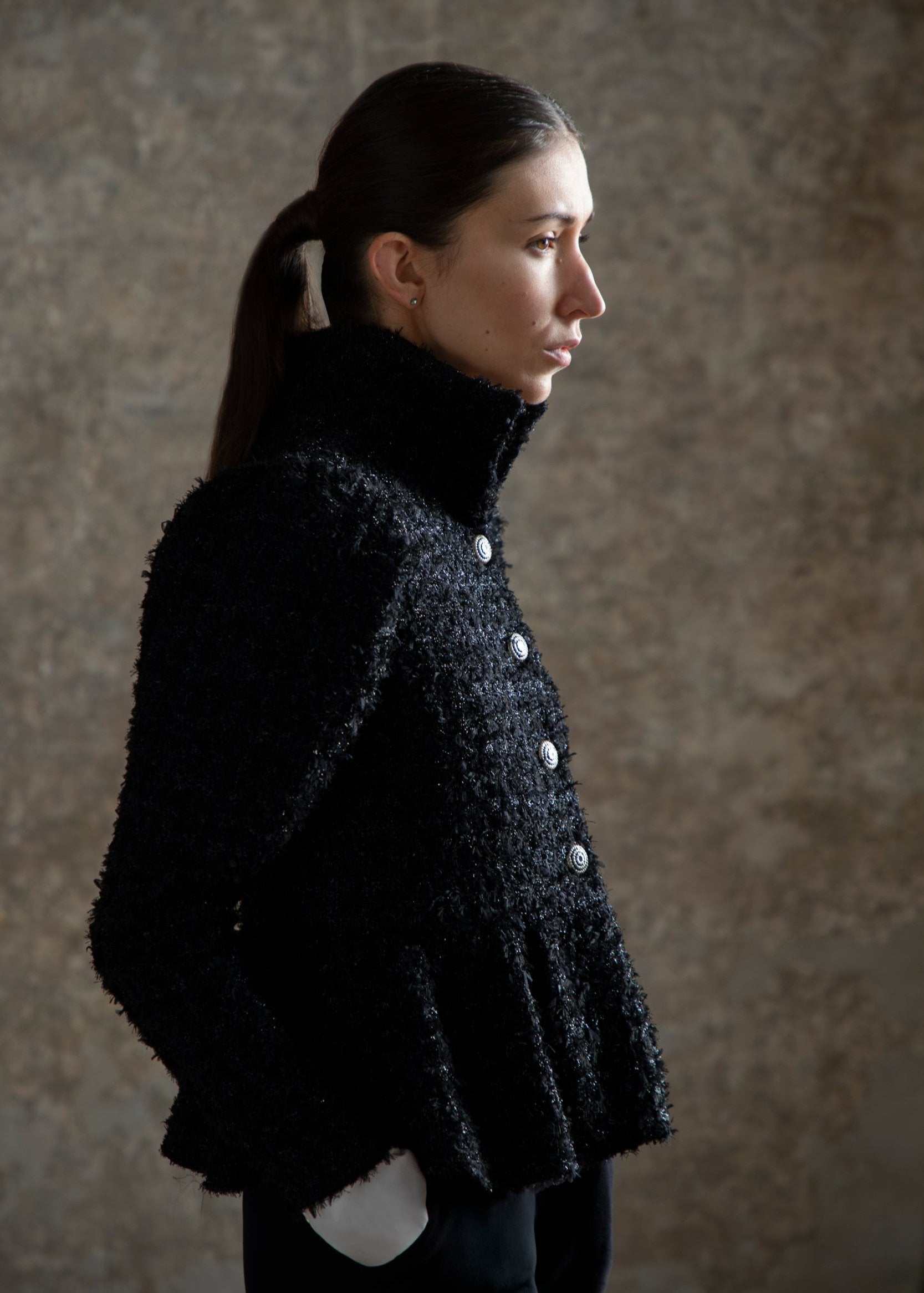Veste en tweed noir luxueuse présentée sur un modèle féminin, vue de profil. La veste arbore un col haut et des boutons décoratifs en métal, tandis que le tissu texturé et épais met en valeur le style chic et sophistiqué. Le modèle a les cheveux attachés et s'affiche dans un décor neutre qui souligne l'élégance de la pièce.