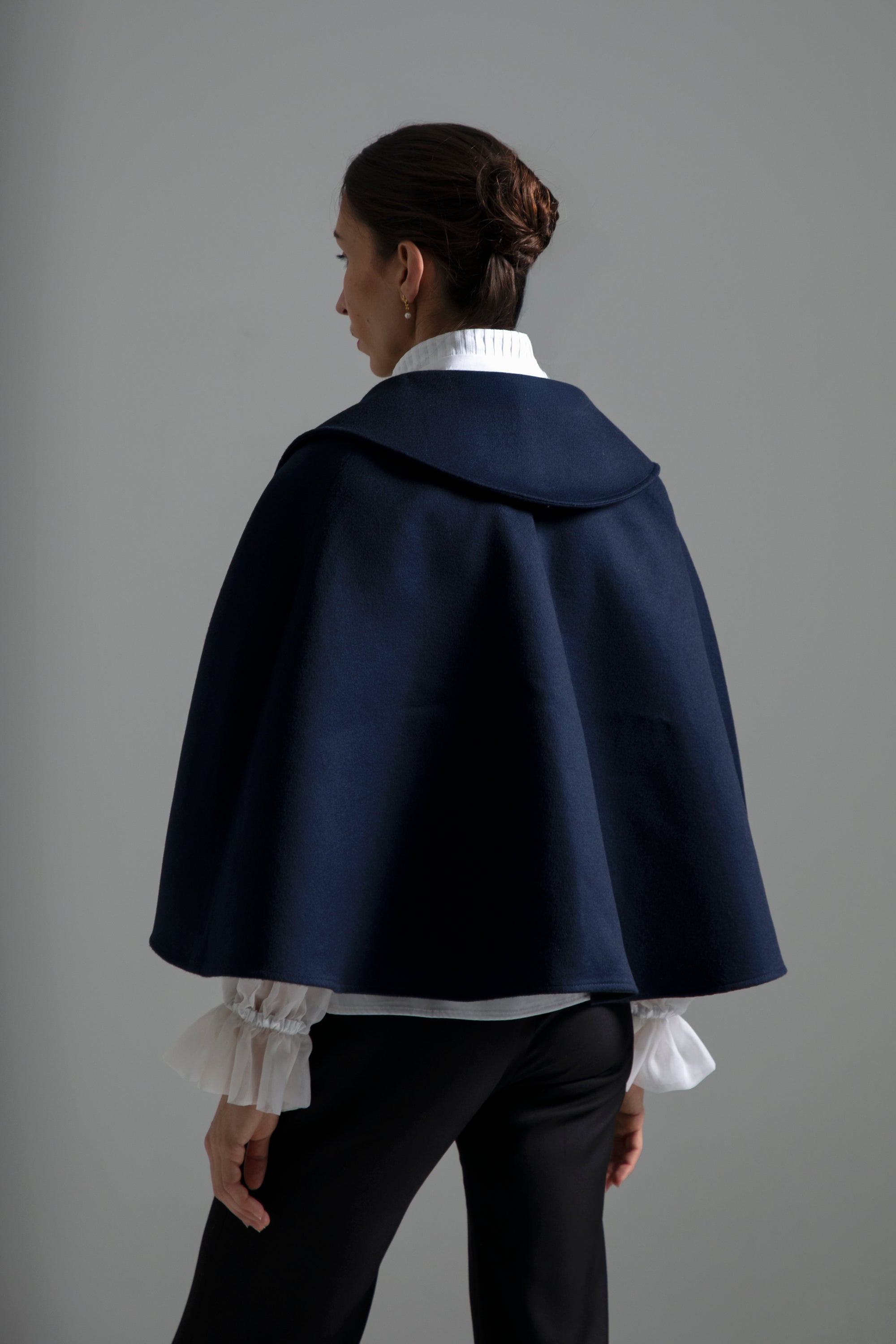 Femme vue de dos, se tenant debout avec le visage légèrement tourné vers la gauche. Elle porte la cape Ondine bleu nuit, en laine et soie, courte à la taille avec des plis en godet dans le dos. Sous la cape, le chemisier Isolde blanc et le pantalon Salomé noir complètent la tenue. 
