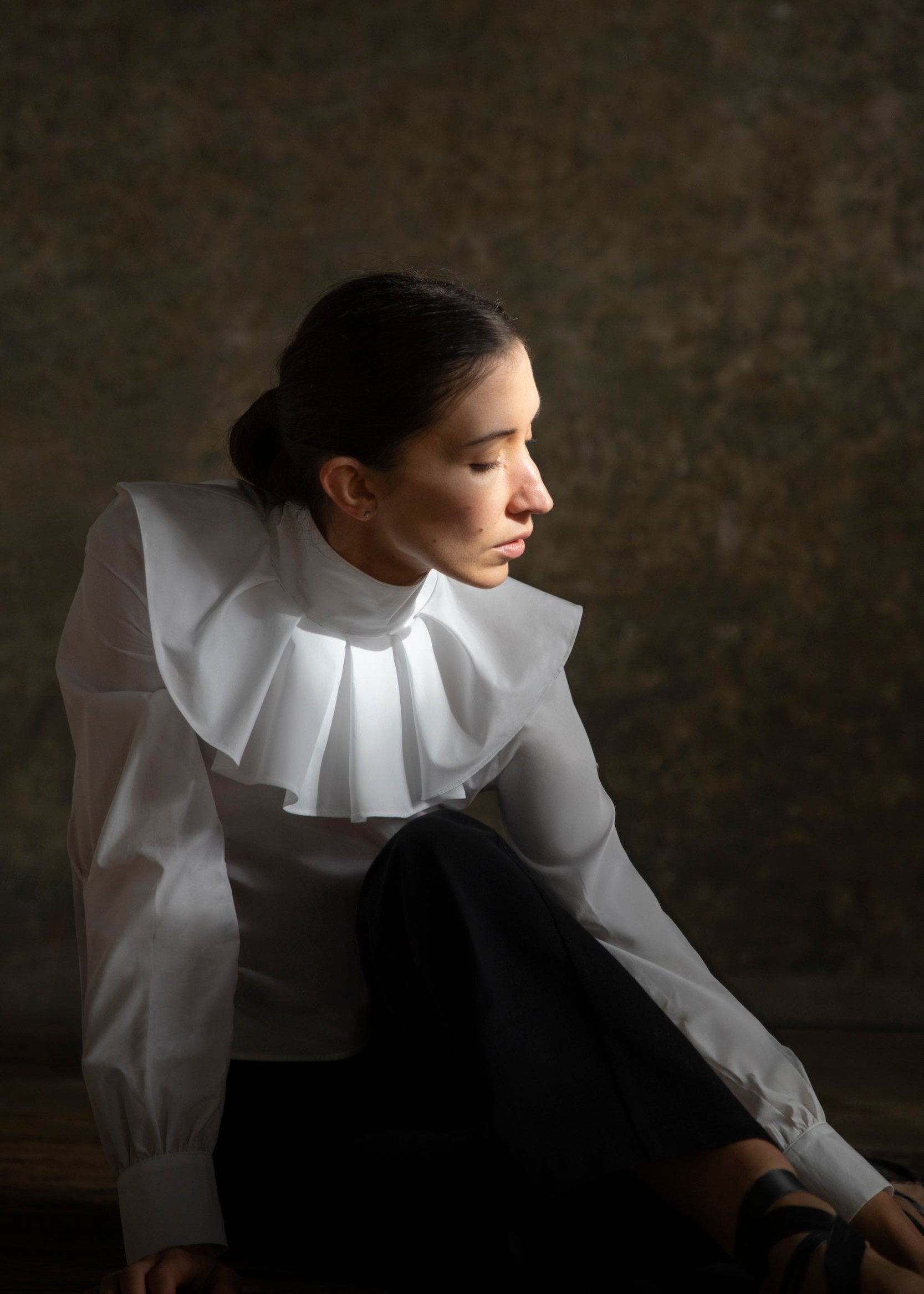 Une femme élégante, vêtue d'une blouse en popeline de coton blanche avec un col large et plissé, se tient assise dans une pose gracieuse. Son regard est tourné vers le bas, exprimant une mélancolie poétique. La lumière douce accentue les détails de la blouse, soulignant la texture et la fluidité du tissu. Le fond flou et sombre met en valeur la délicatesse de sa silhouette, évoquant une atmosphère de ballet et de grâce.