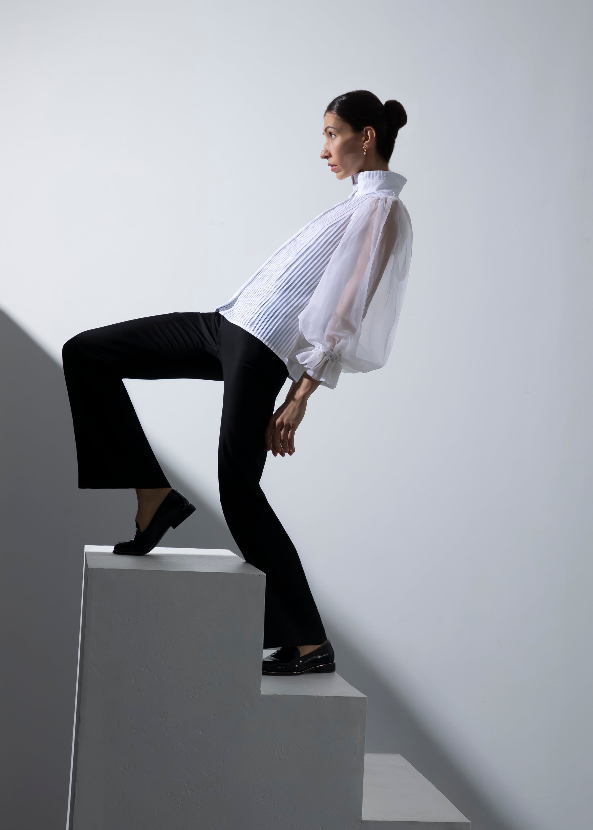 Femme montant les marches de manière artistique en inclinant son corps vers l'arrière. Elle porte le chemisier Isolde et le pantalon Salomé pour un contraste entre ombre et lumière.
