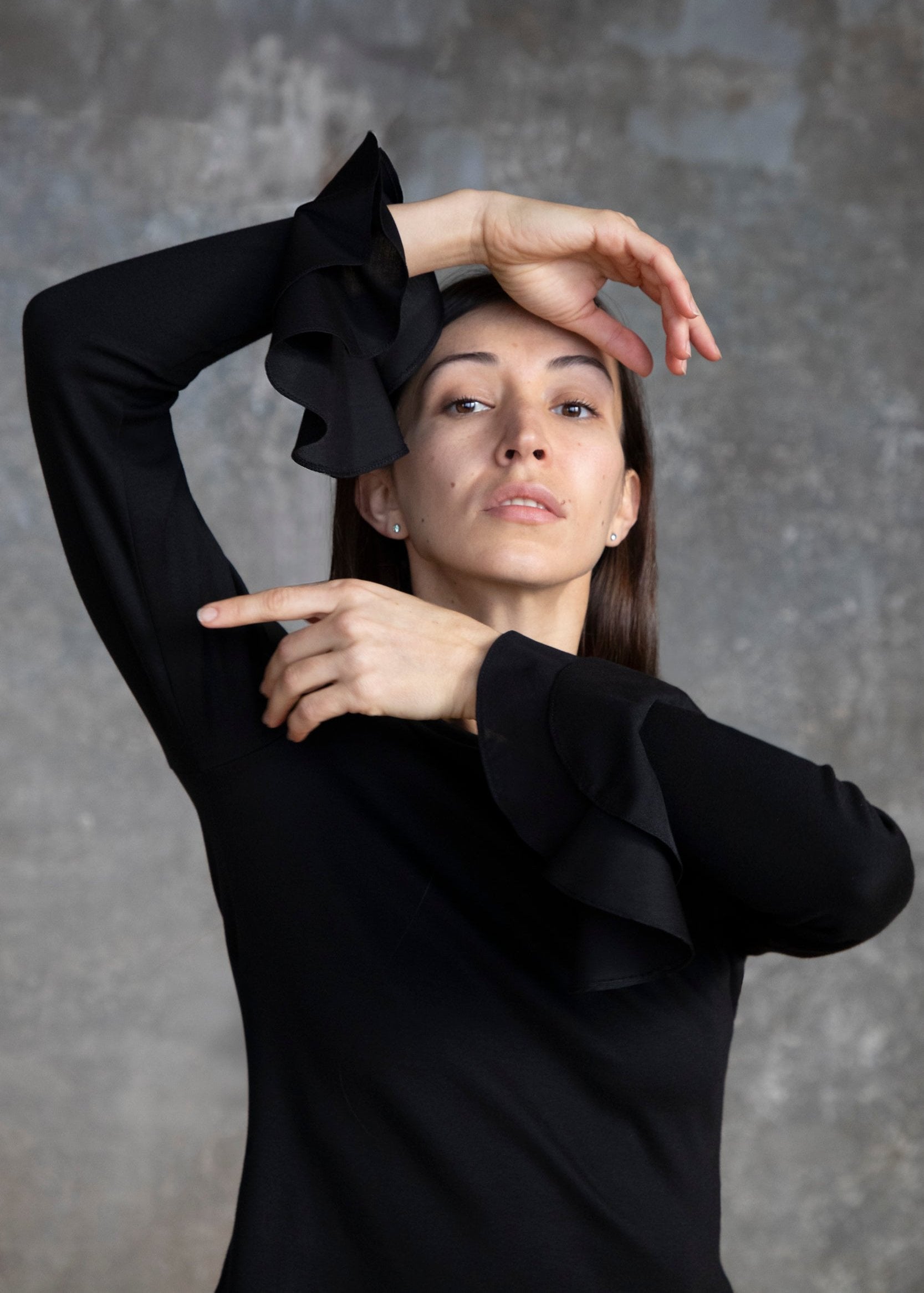Une femme pose en portant un t-shirt noir à manches longues, orné de volants élégants au niveau des poignets. Elle se tient droite, la tête légèrement inclinée vers l'avant et le bras gauche levé, la main posée sur son front. Son expression est confiante et sophistiquée, mettant en valeur l'élégance du vêtement. L'arrière-plan est flou et neutre, ce qui accentue le t-shirt.