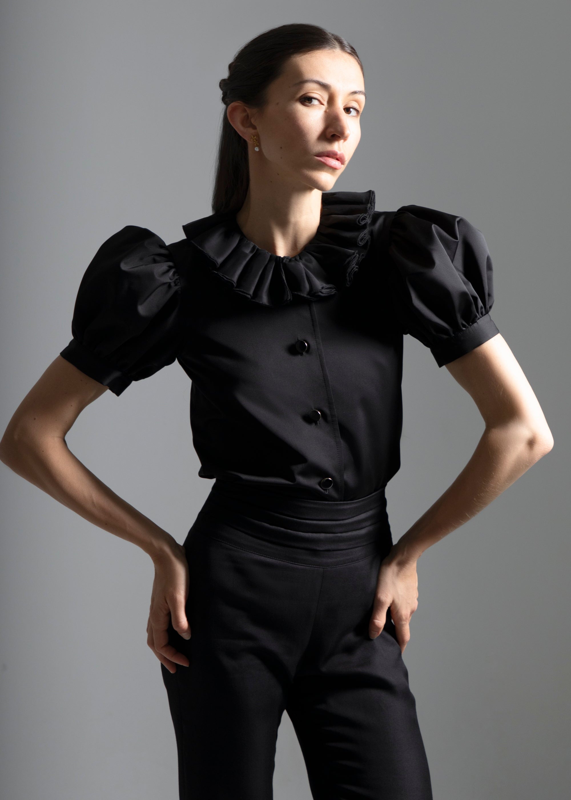 Une femme habillée en Sidonie Paris, portant un pantalon noir taille haute avec une ceinture plissée et un top avec des manches ballons et un col en volants, se tient debout avec un léger sourire et les mains sur les hanches.