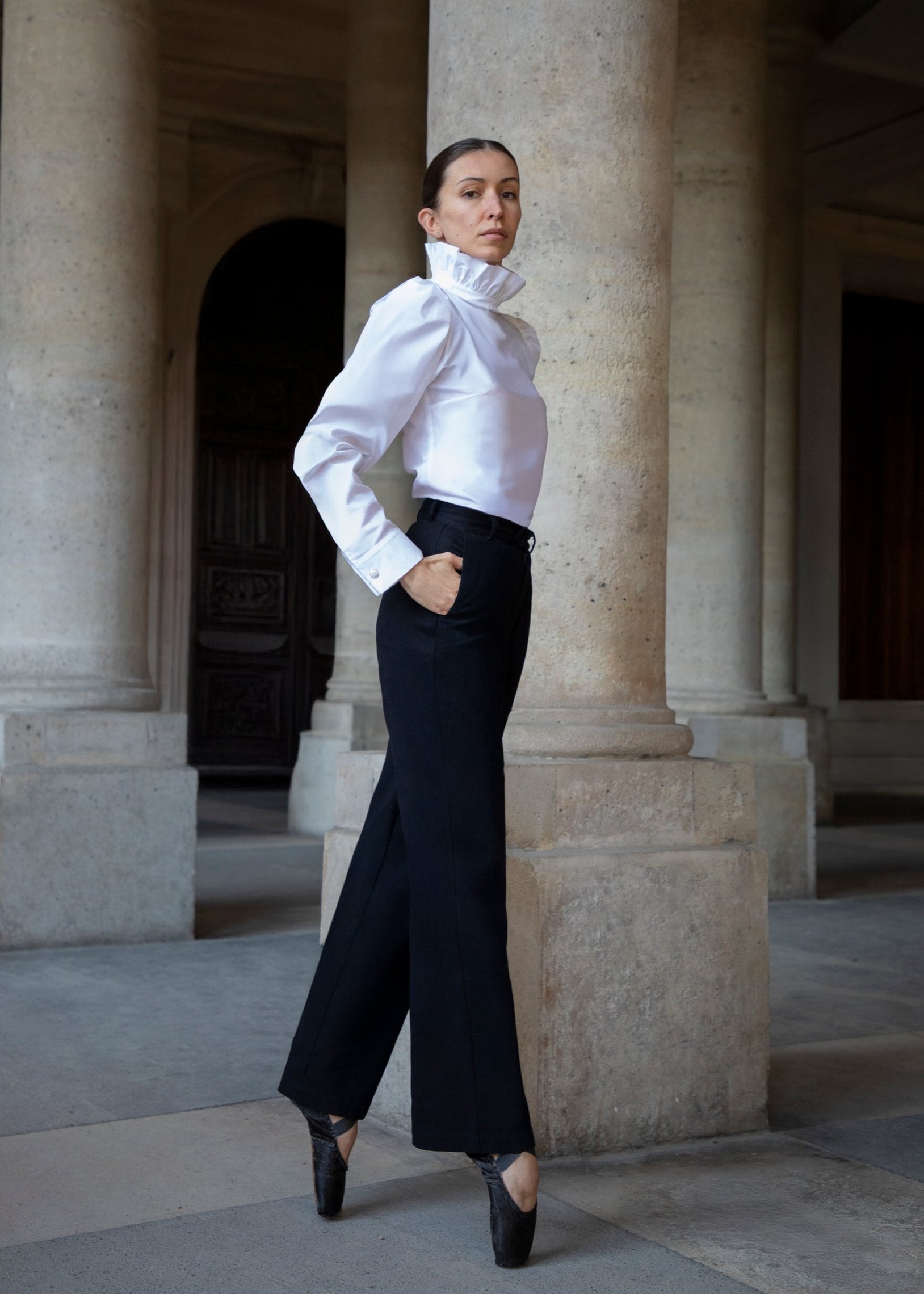 Une femme vêtue d'une chemise Sidonie blanche et d'un pantalon noir Raymonda, se tient sur ses pointes de danse devant de hautes colonnes de pierre. Ses cheveux sont tirés en arrière et ses mains sont dans ses poches, dégageant une impression d'élégance et de force.