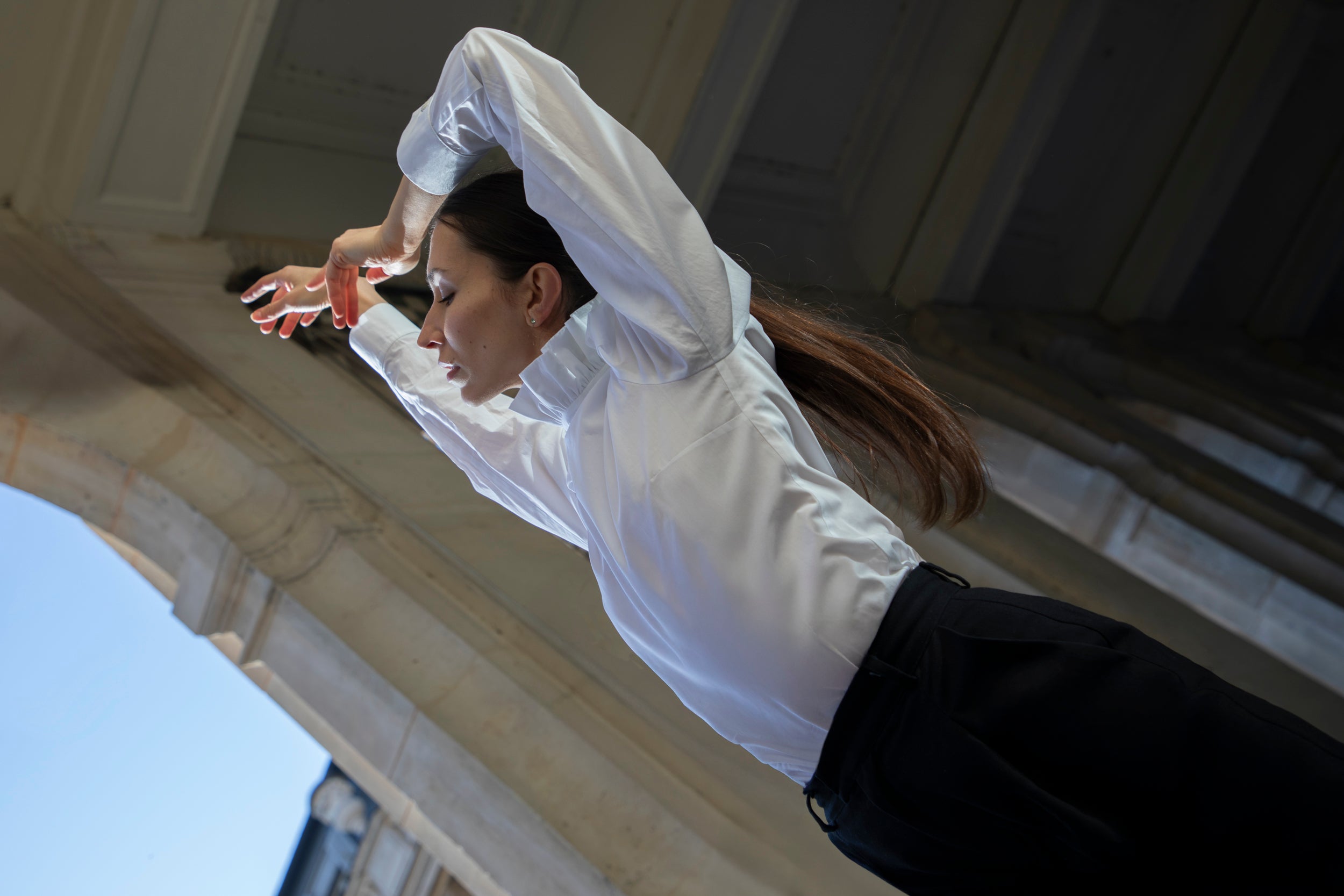 Vêtue de Sidonie Paris, une danseuse exécute une pose gracieuse sous une arche de pierre, dégageant une élégance avec les bras gracieusement courbés au-dessus de sa tête, sur fond de colonnes architecturales et de ciel bleu.