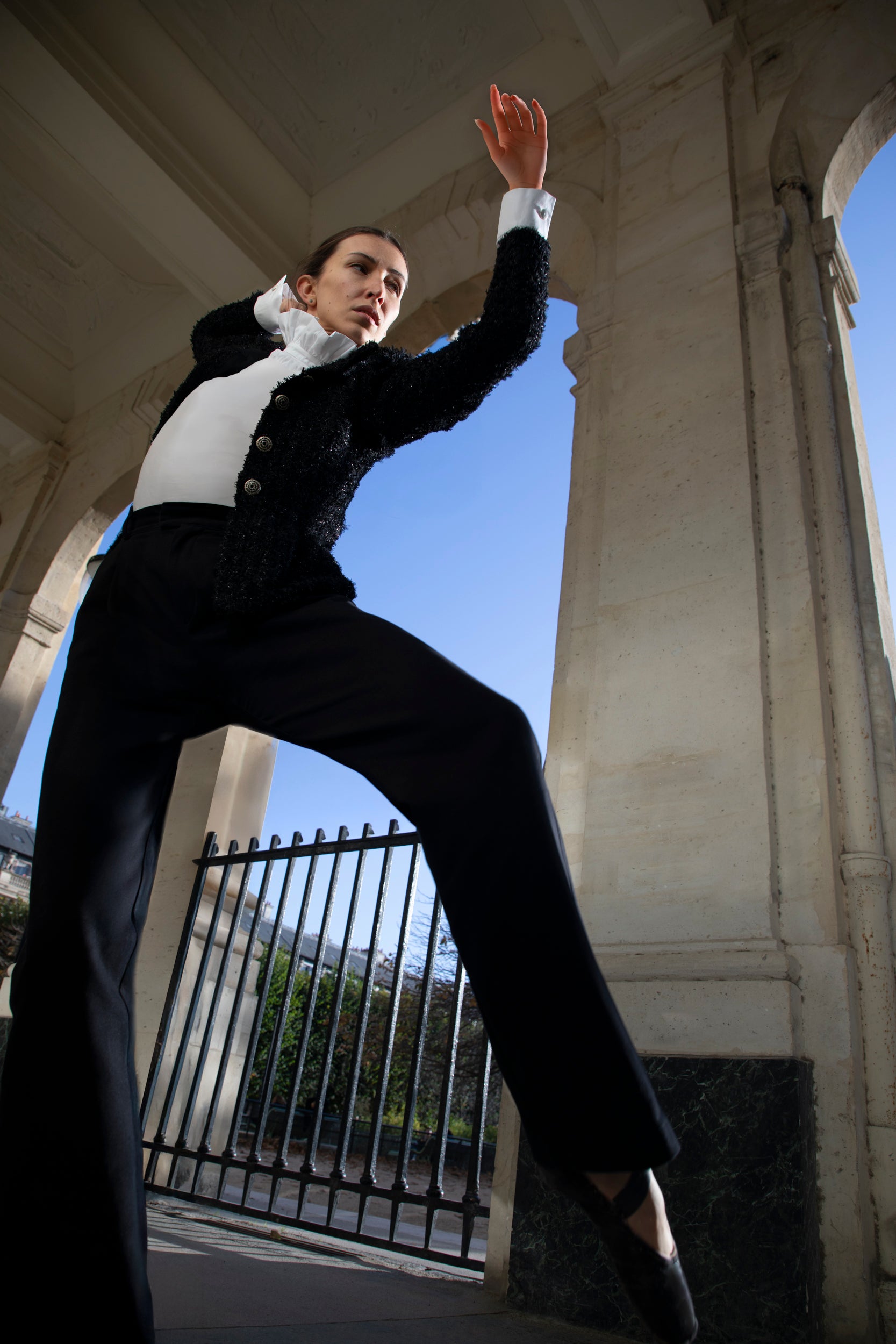 Une personne vêtue en tenue Sidonie Paris, composée d'une veste en tweed et d'un pantalon noirs, pose de manière dynamique sous une arche de pierre sur fond de ciel bleu clair. La prise de vue en contre-plongée met en valeur le mouvement gracieux et la posture pleine d'assurance.