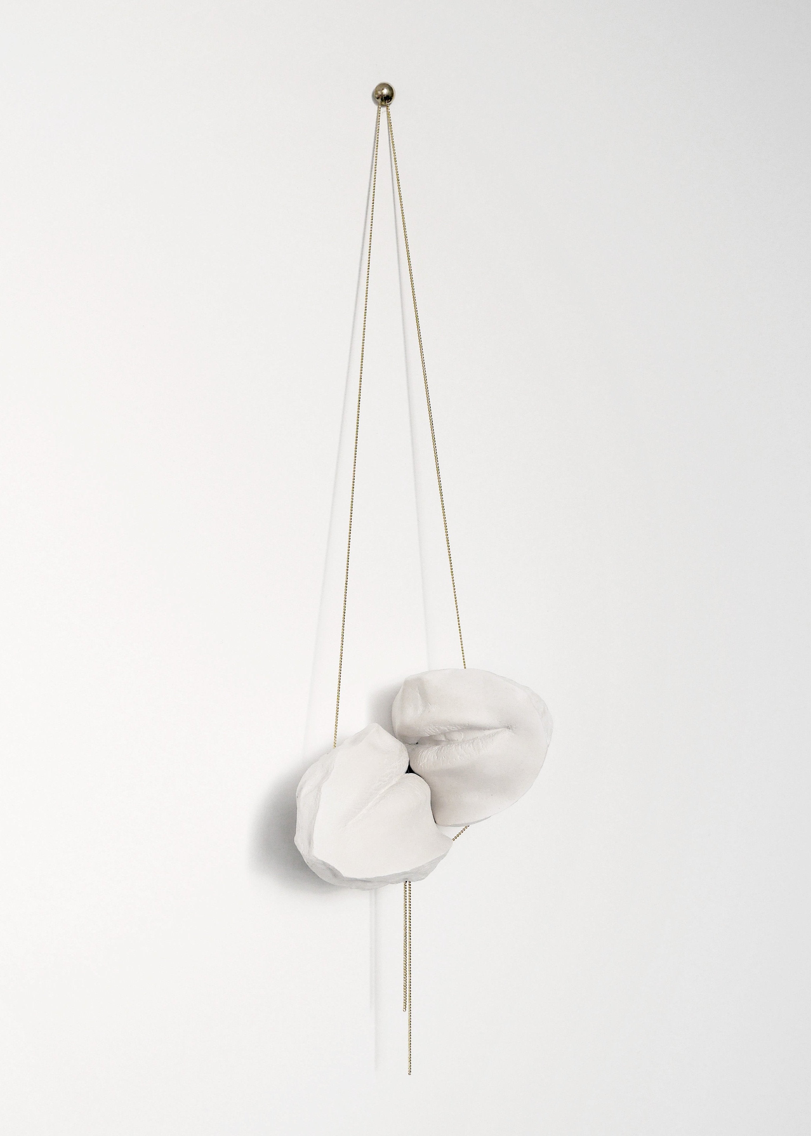 Sculpture en plâtre suspendue, composée de deux bouches blanches, accrochée à un mur par des fils fins dorés, avec un support en haut. L'œuvre d'art évoque une esthétique moderne et minimaliste, jouant sur la légèreté et la texture du matériau.