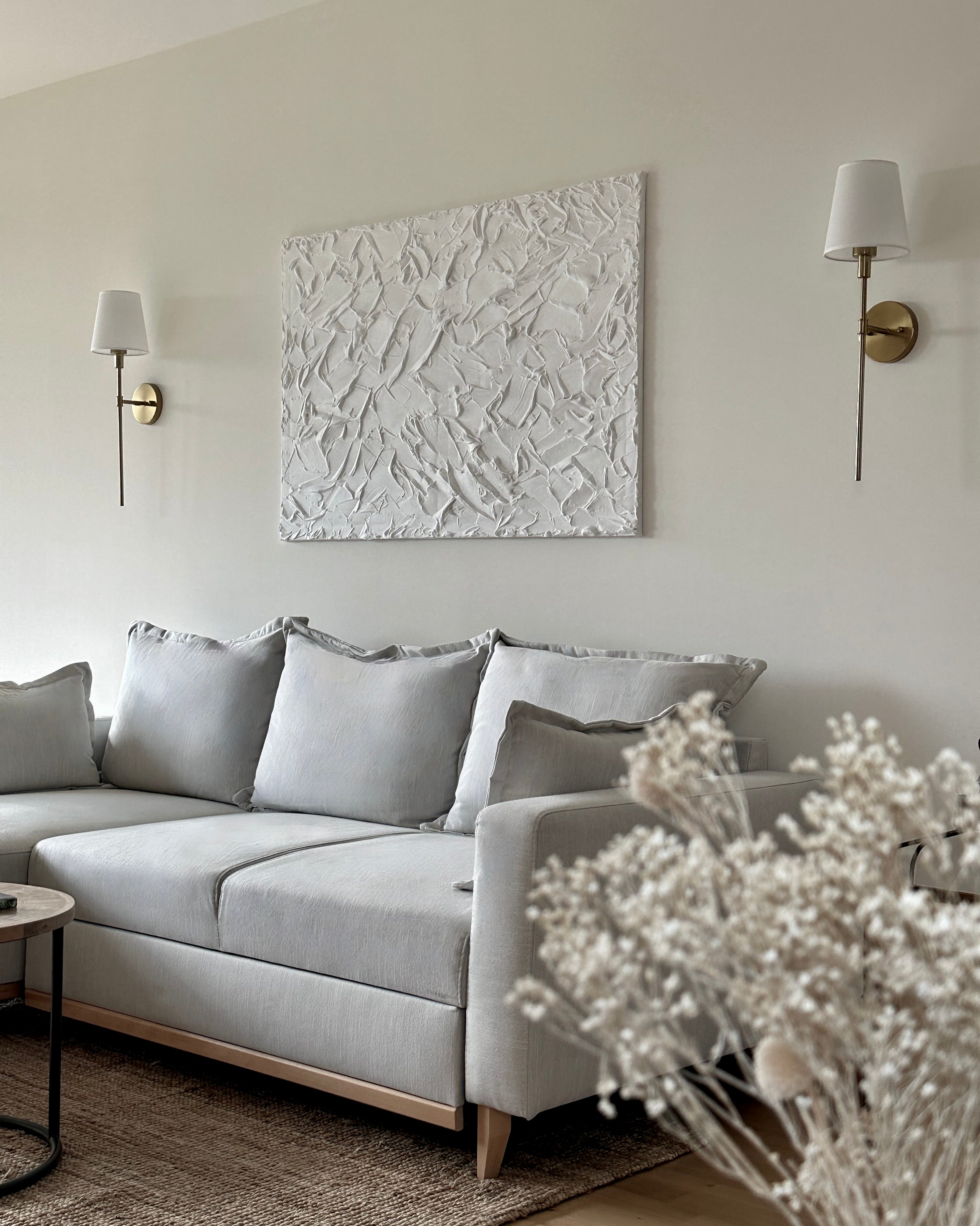 Tableau texturé en relief accroché au mur d'un salon moderne, surplombant un canapé beige clair avec des coussins assortis. Le mur est peint en blanc gris, et des appliques murales dorées ajoutent une touche d'élégance. Une petit table basse en bois et métal se trouve devant le canapé, tandis qu'un bouquet de fleurs séchées apporte une ambiance chaleureuse à la pièce.